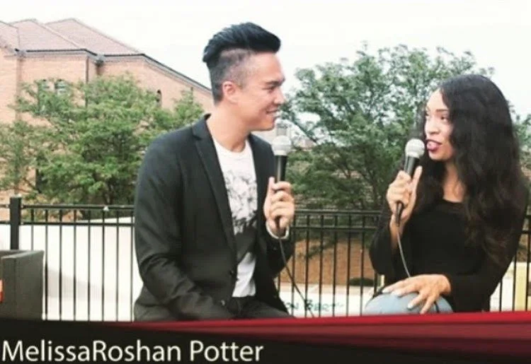 Tom McClur and MelRo sitting outside, holding microphones, engaged in conversation during an interview or discussion, with trees and a brick building in the background.