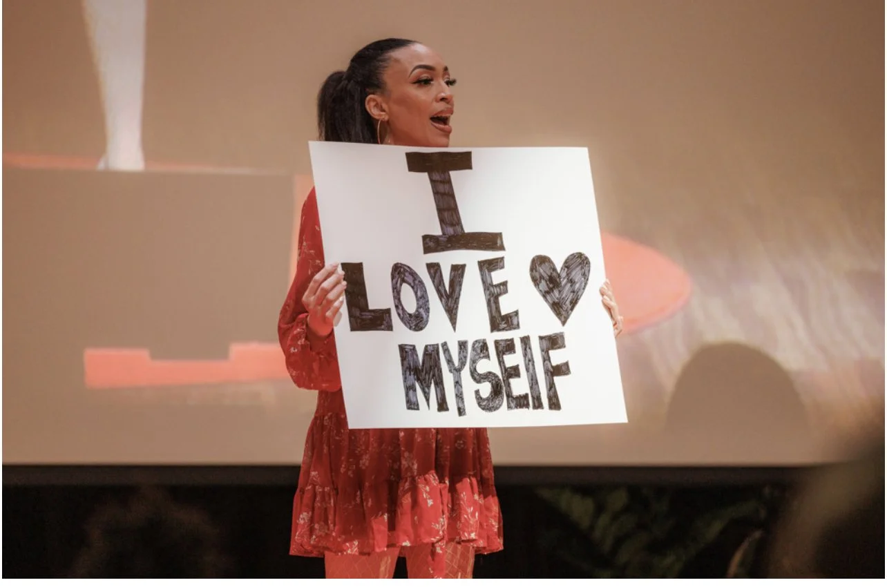 MelRo holding a sign that says, 'I love myself' with a heart symbol, standing on stage.