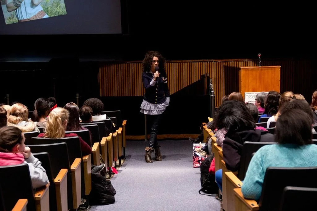 MelRo standing on a stage speaking into a microphone to an audience seated in an auditorium.