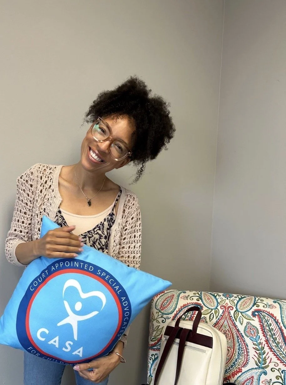 MelRo with glasses and curly hair smiling and holding a blue pillow with the CASA logo, standing next to a patterned couch and a white bag.
