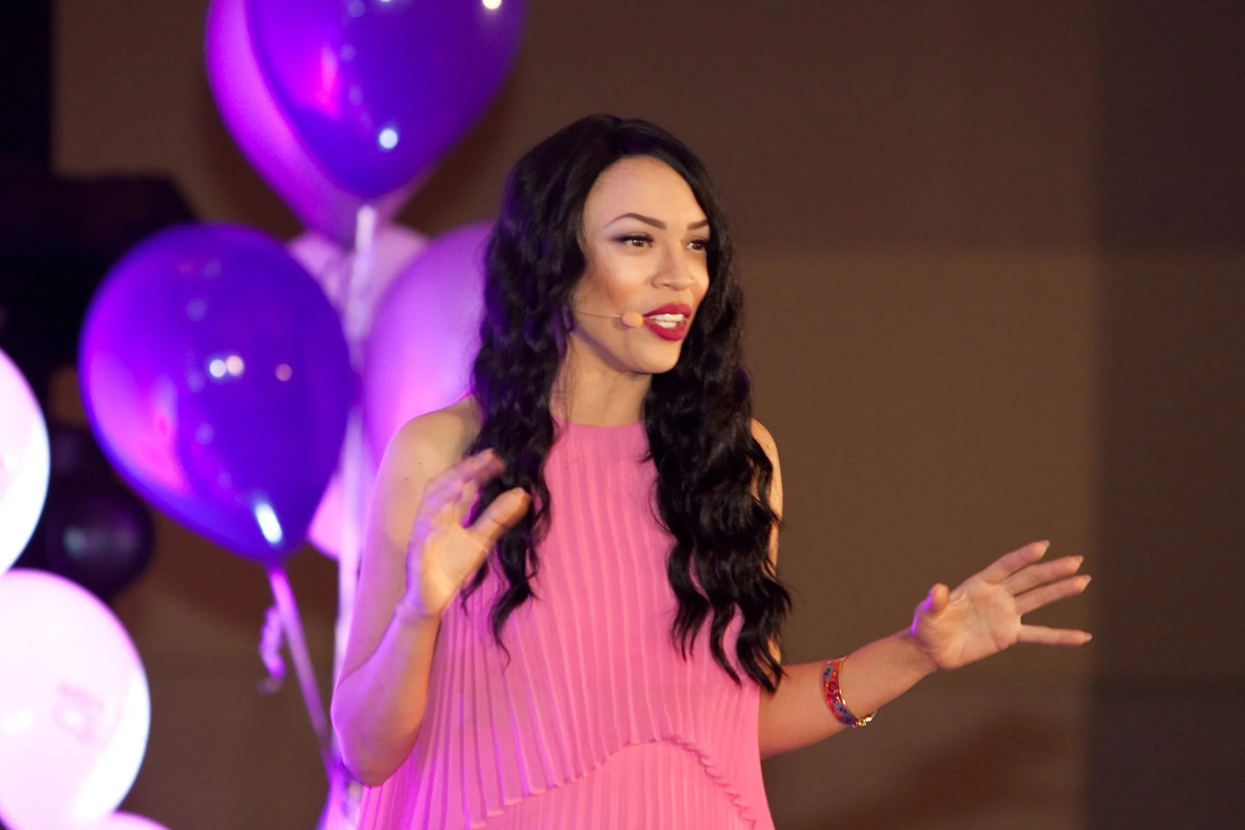 MelRo with long dark wavy hair, wearing a pink dress and a red bracelet, speaking into a headset microphone with purple balloons in the background.