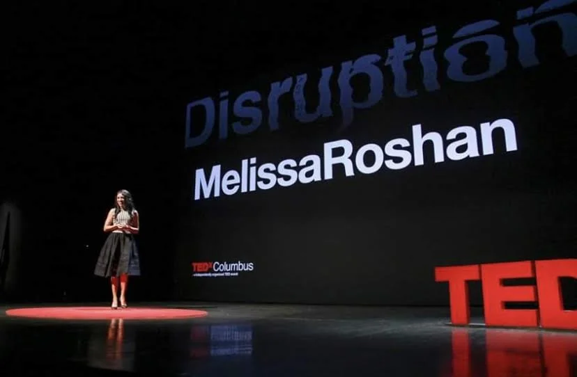 A woman standing on a stage next to a large screen displaying her name, Melissa Roshan, at a TEDx event, with red TED letters on the floor.
