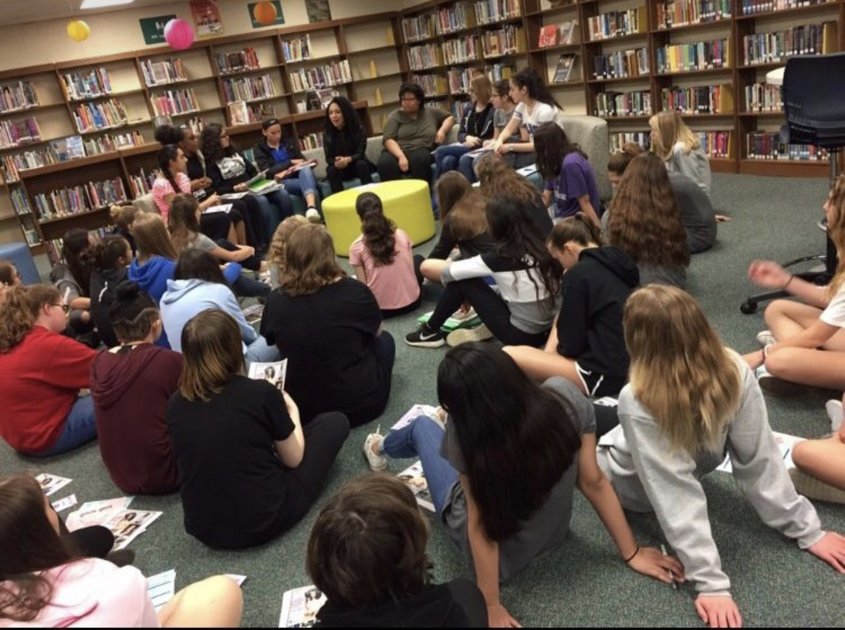 Group of students sitting and listening to a woman speaking in a library.