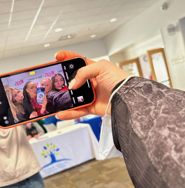 Person taking a photo of four women at an event, with a backdrop displaying a pink logo and a table with a blue logo in the background.