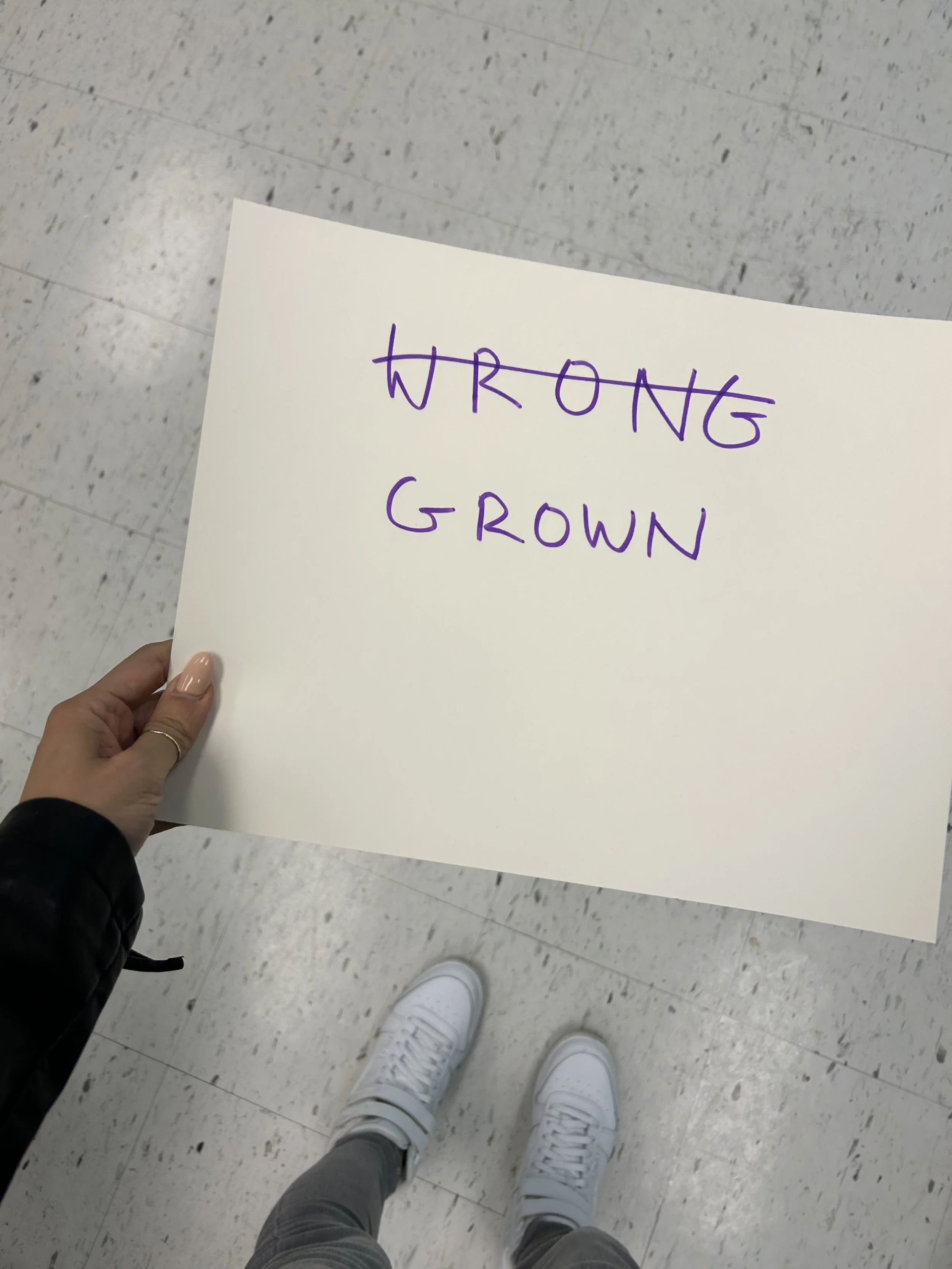 Person holding a sign with the words 'WRONG GROWN', with 'WRONG' crossed out, in purple ink, standing on a shiny tiled floor.