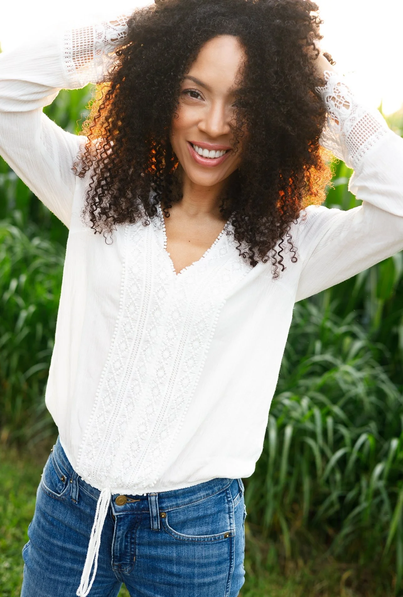 MelRo with curly hair smiling brightly in a white embroidered blouse and blue jeans, standing outdoors with green foliage in the background.