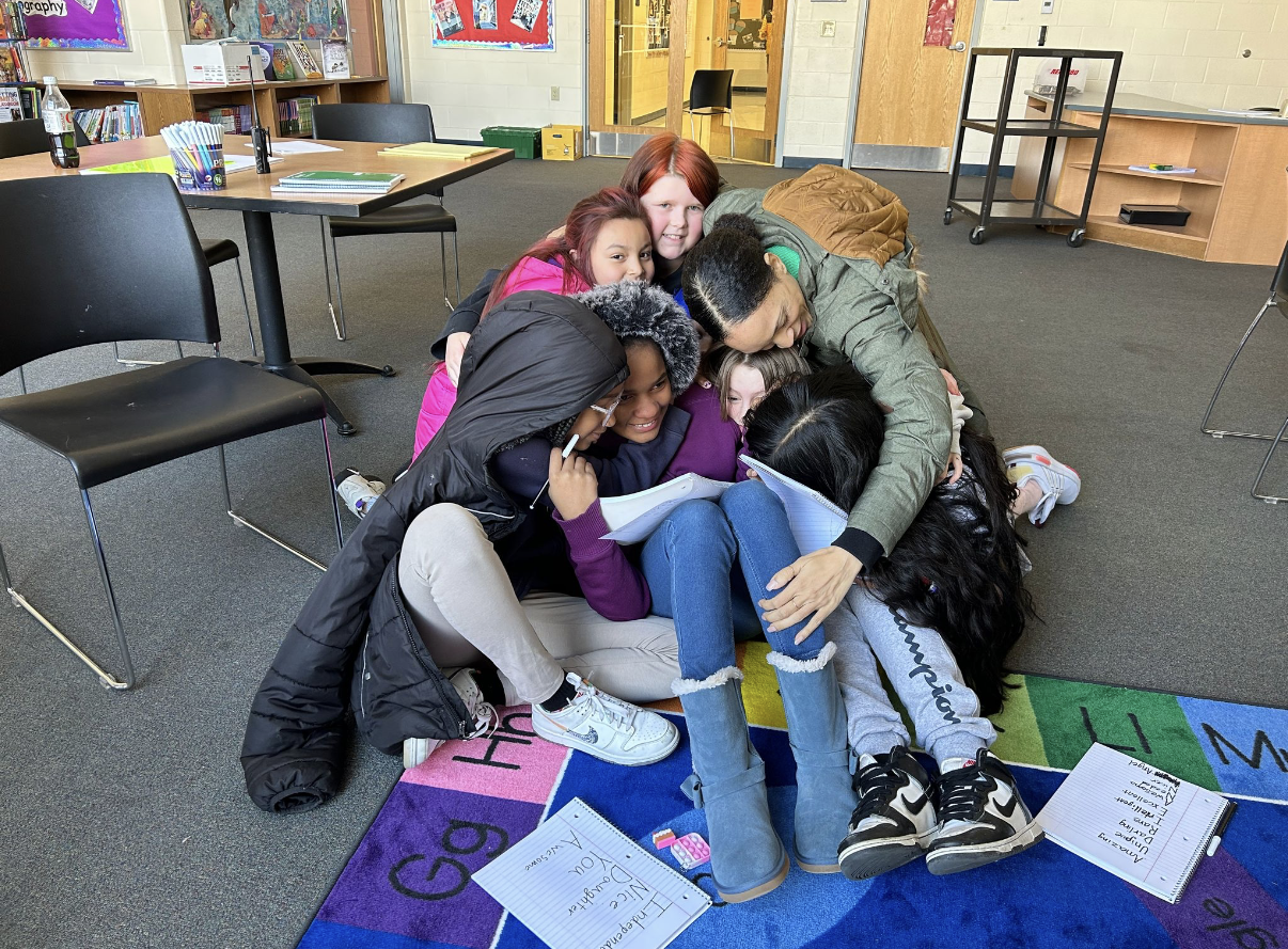 A group of children and an adult are gathered together on the floor of a classroom, hugging and looking at a notebook. The classroom has tables, chairs, books, and supplies.