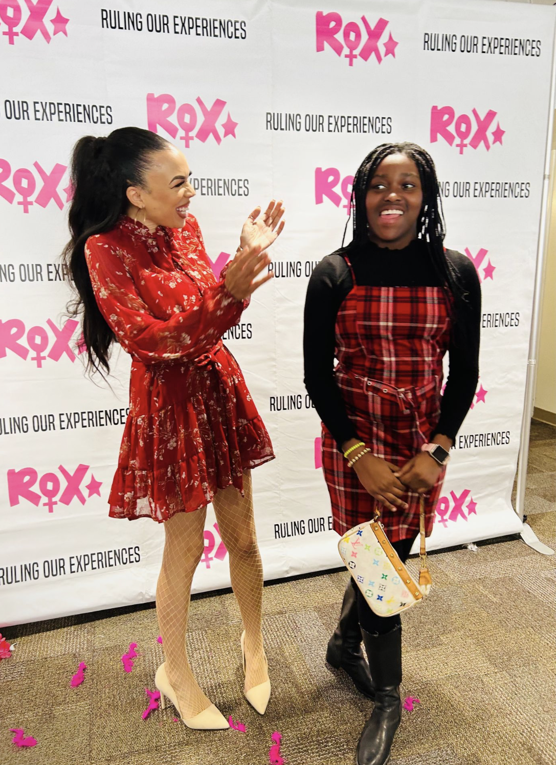Two women standing in front of a backdrop with pink and black text that says 'ROX', 'RULING OUR EXPERIENCES'. The woman on the left, with long dark hair tied in a high ponytail, wears a red floral dress with sheer long sleeves, fishnet stockings, and beige pointed heels, and appears to be smiling and gesturing. The woman on the right, with long braided hair, wears a black and red plaid dress over a black long-sleeve top, black boots, and carries a colorful designer purse. Pink confetti is scattered on the floor.