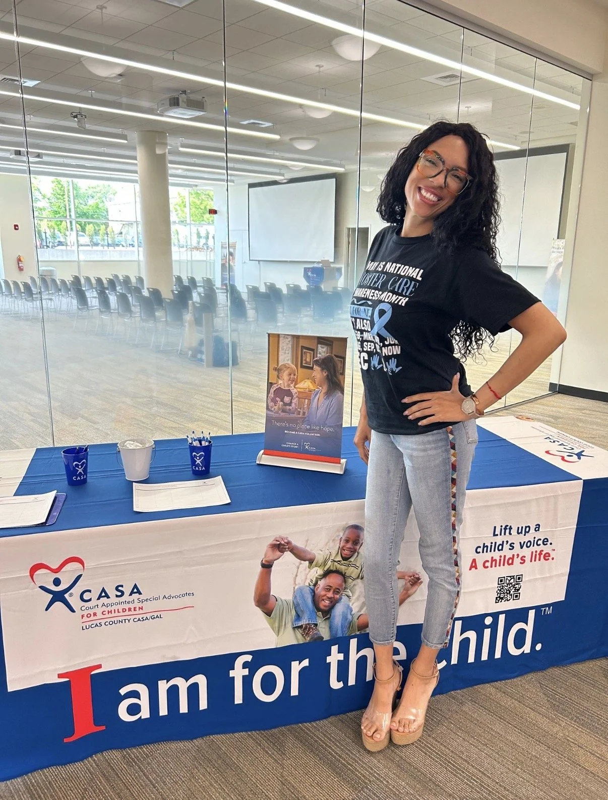 MelRo with black hair, glasses, and a black t-shirt stands smiling next to a table with CASA for Children promotional materials, inside a modern conference room with glass walls and an empty room with chairs in the background.