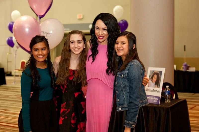 MelRo in a pink dress standing with three young girls inside a decorated event space with balloons and a table with photo display and drinks.