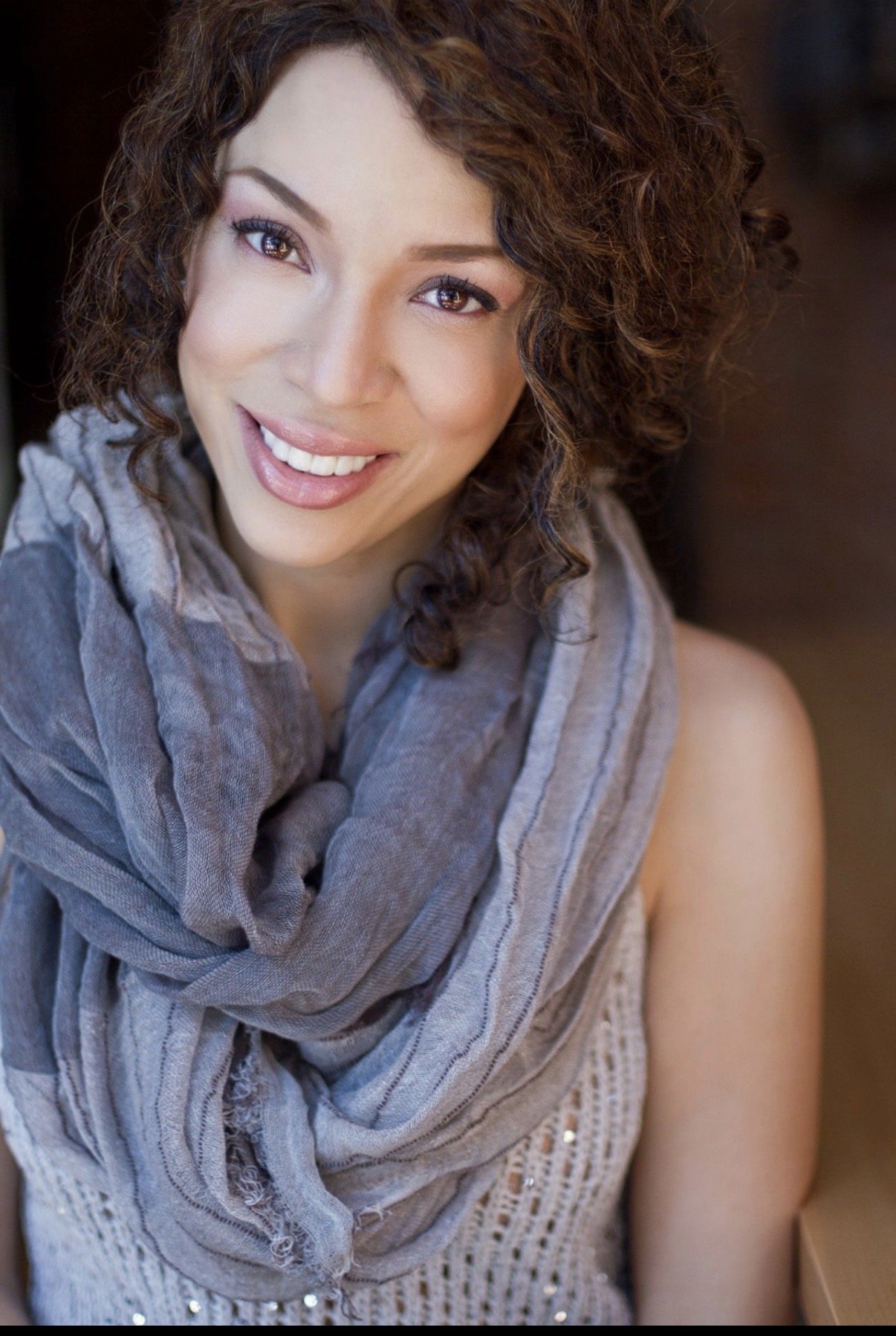 MelRo with curly brown hair smiling at the camera, wearing a gray scarf and a sleeveless top with sequins.