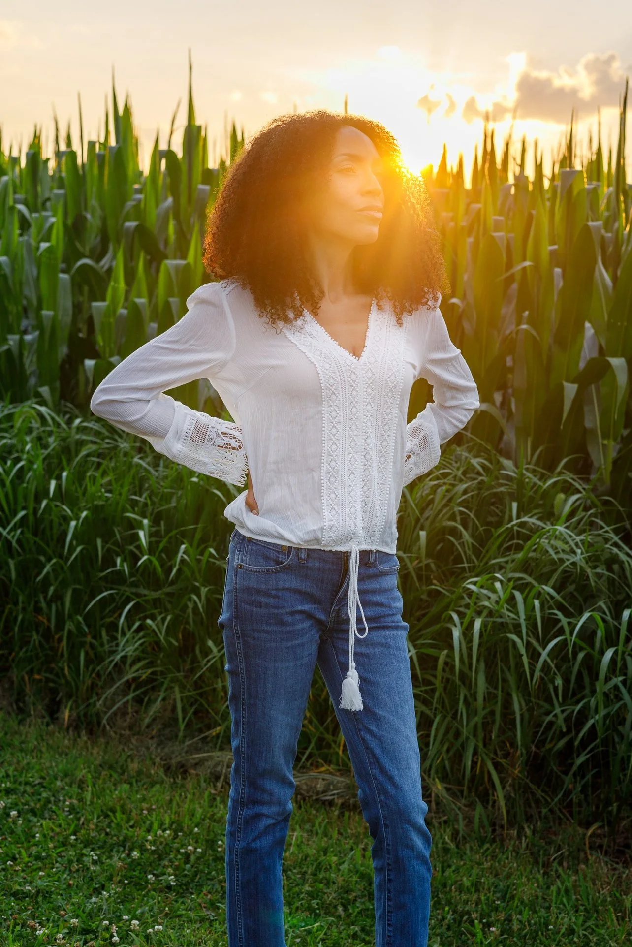 Melissa Roshan stands in front of a cornfield during sunset, wearing a white blouse and blue jeans, with hands on hips.