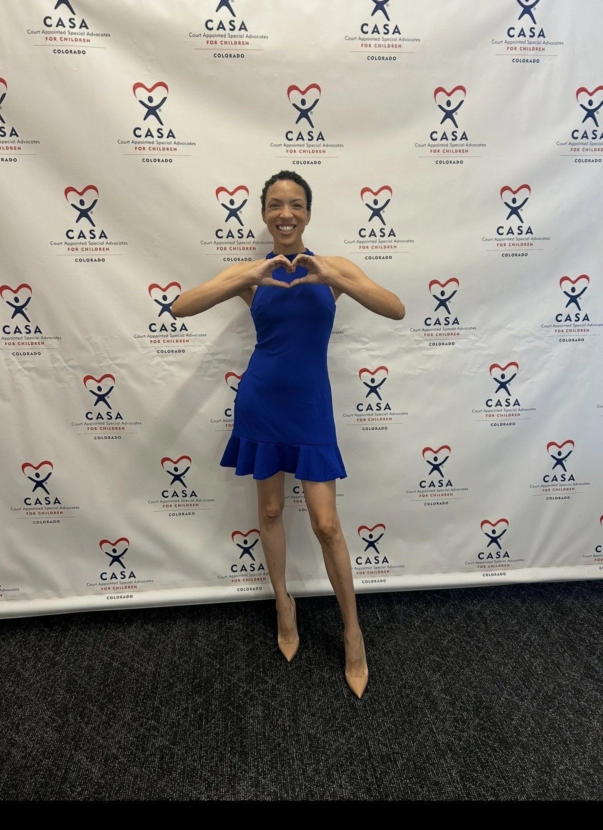 MelRo in a blue dress making a heart shape with her hands in front of a step-and-repeat backdrop with "CASA Colorado" and heart-themed logos.