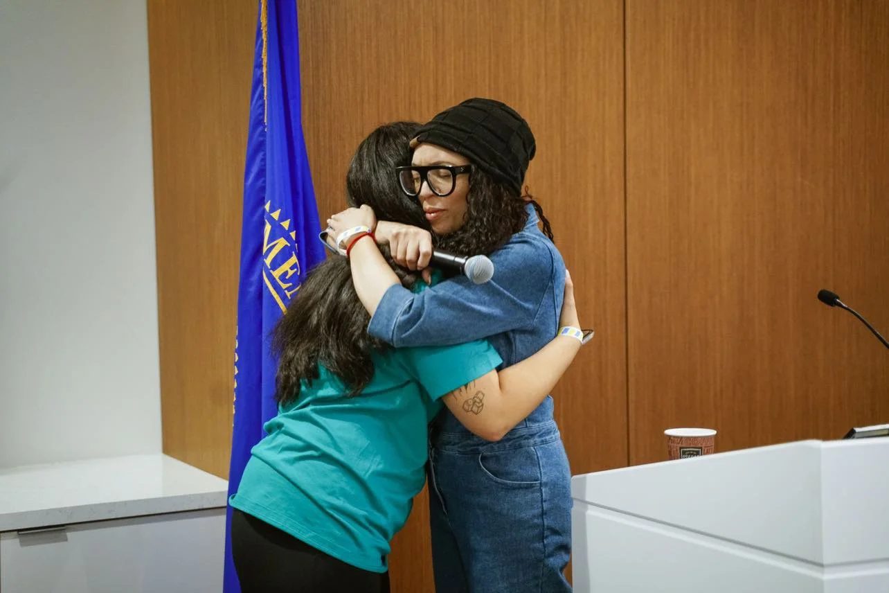 MelRo hugging a student in an indoor setting with a wooden wall, a blue flag, and a table with a coffee cup, microphone, and podium.
