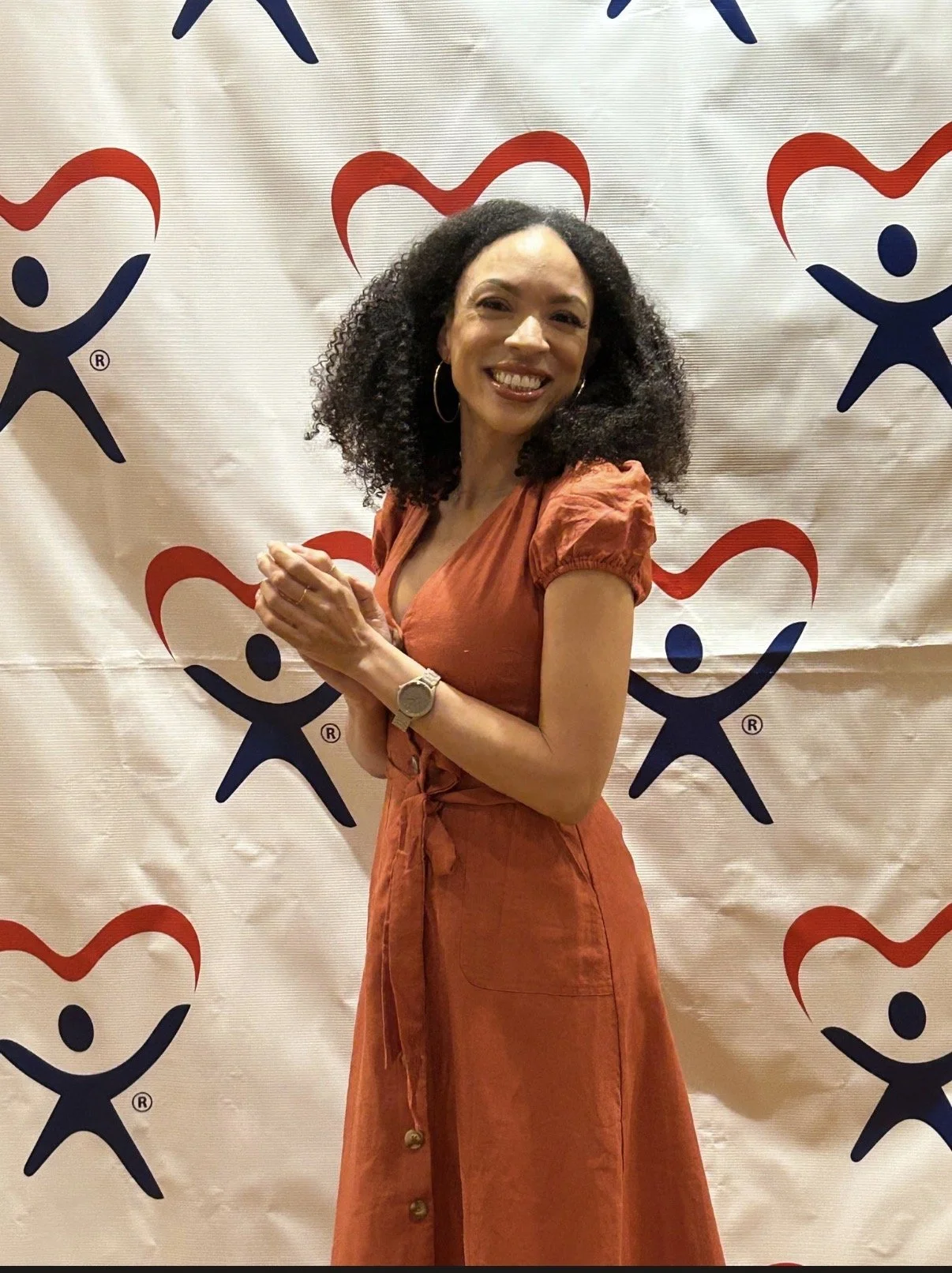 MelRo wearing an orange dress, smiling, standing in front of a backdrop with multiple blue and red hearts and figures of people.