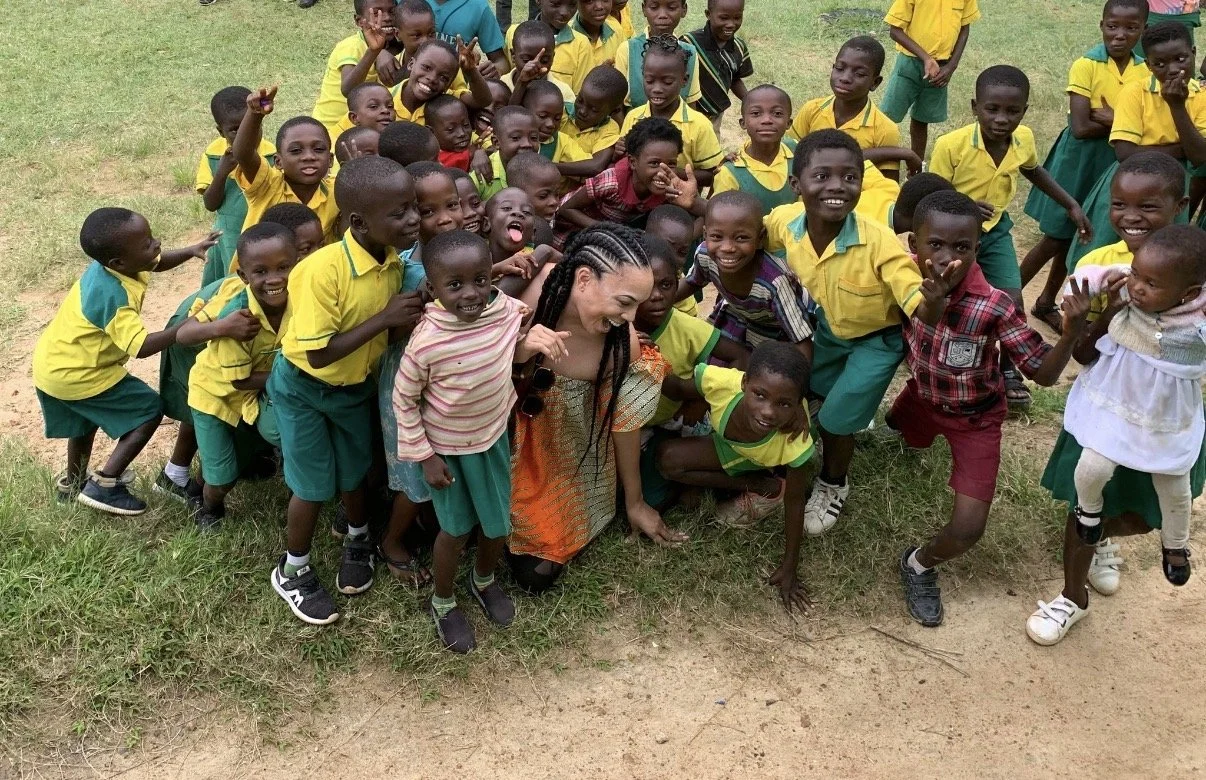 A group of children in school uniforms, along with a MelRo, are gathered outdoors on a grassy area, smiling and posing for the camera.