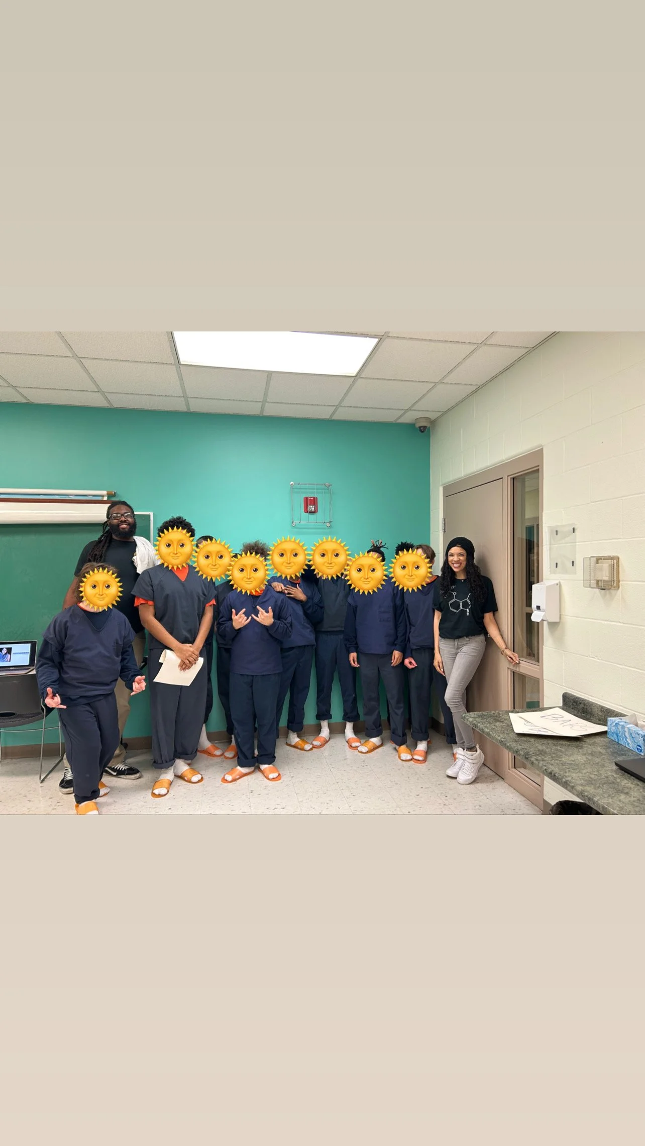 Group of students and two teachers in a classroom, with sun emoji face filters over their faces, all wearing matching orange slippers, posing for a photo.