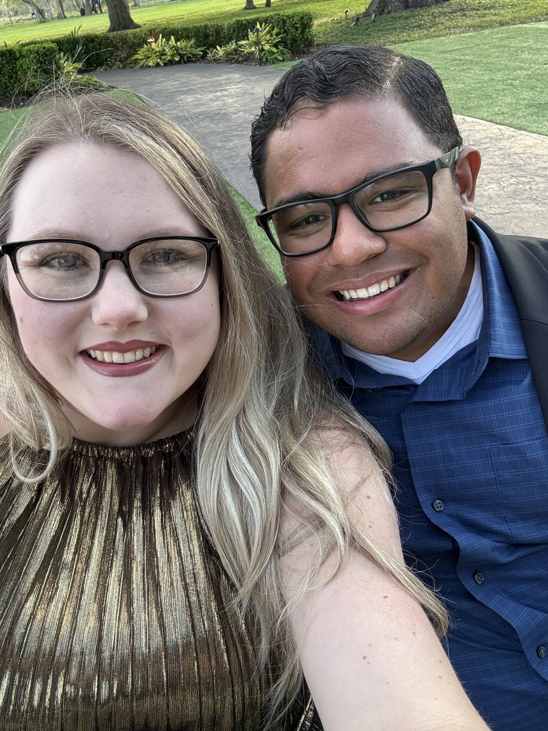 A close-up selfie of a woman and a man outdoors in a park with green grass, trees, and a pathway in the background.