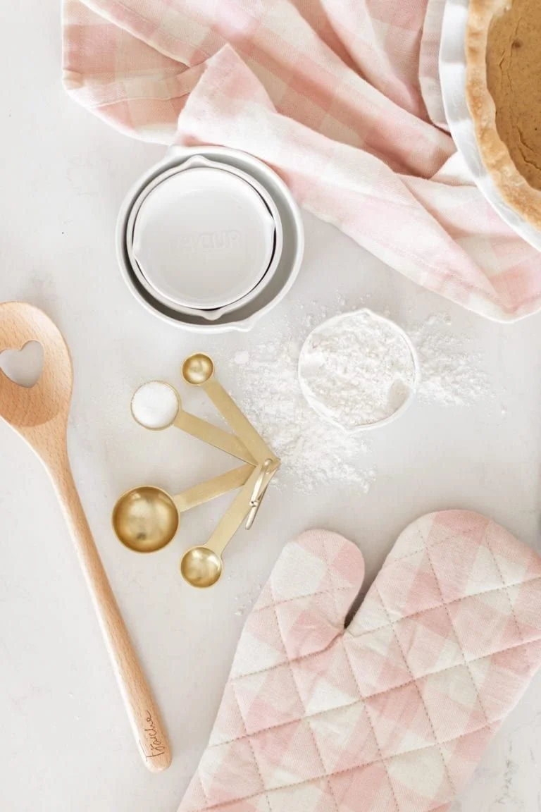 Baking ingredients and tools on a white surface, including a wooden spoon, a container of flour, a piping tip, gold and white measuring spoons, pink oven mitts, and a piece of pink and white checkered cloth.