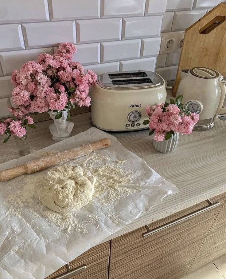A kitchen countertop with a ball of dough, a rolling pin, and flour on parchment paper, flanked by pink flowers in vases. There is a cream-colored toaster and kettle, a cutting board, and white wall tiles in the background.