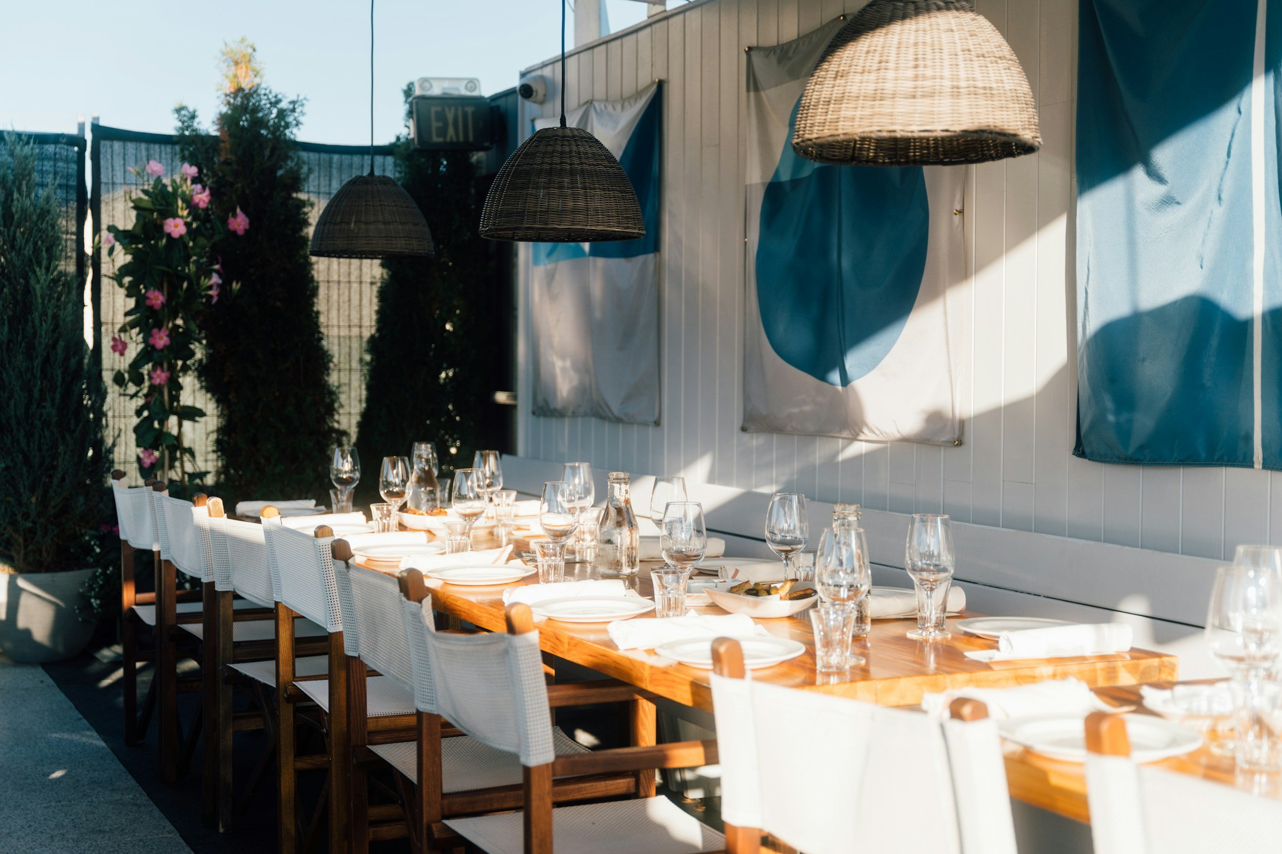 Outdoor dining table set with white plates, glasses, and napkins, illuminated by hanging wicker lamps, on a patio with decorative wall art and plants in the background.