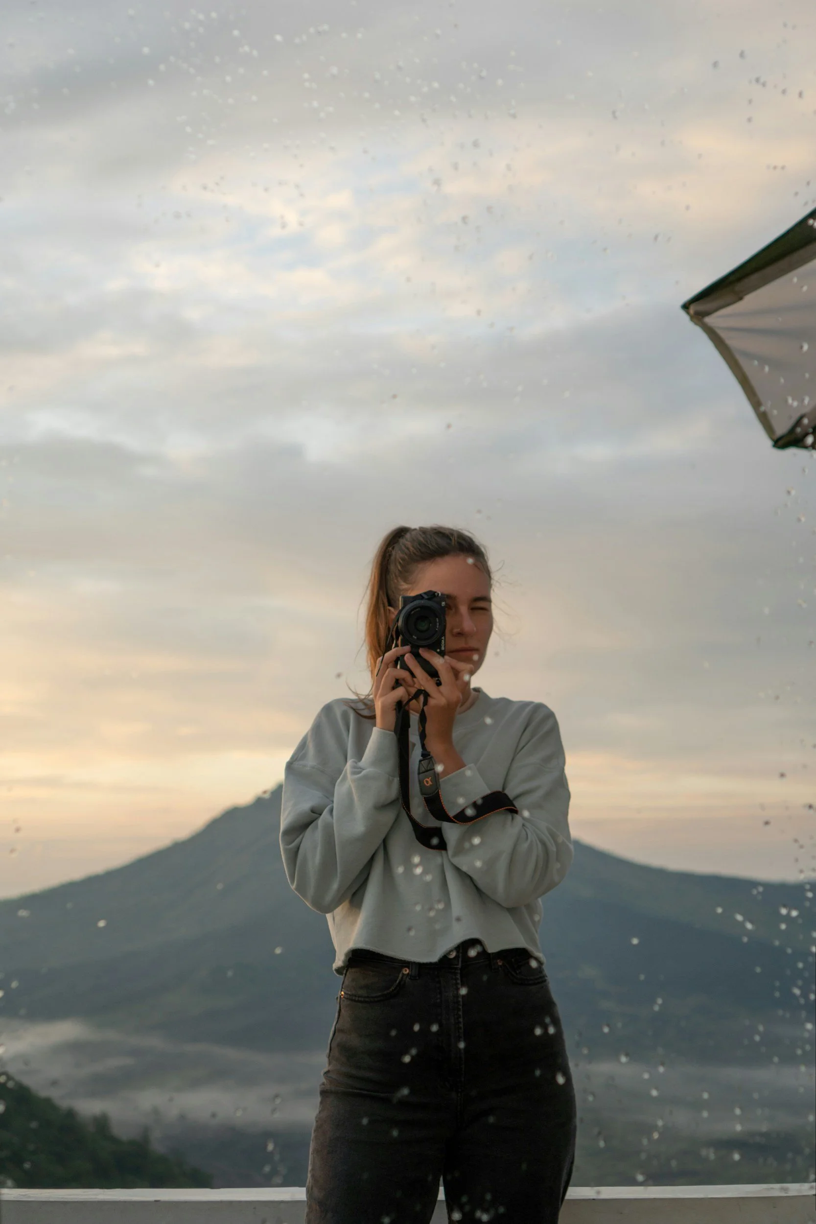 A woman with long hair tied in a ponytail taking a photo of herself with a camera in front of a mountain landscape with cloudy sky and rain droplets on a glass surface.