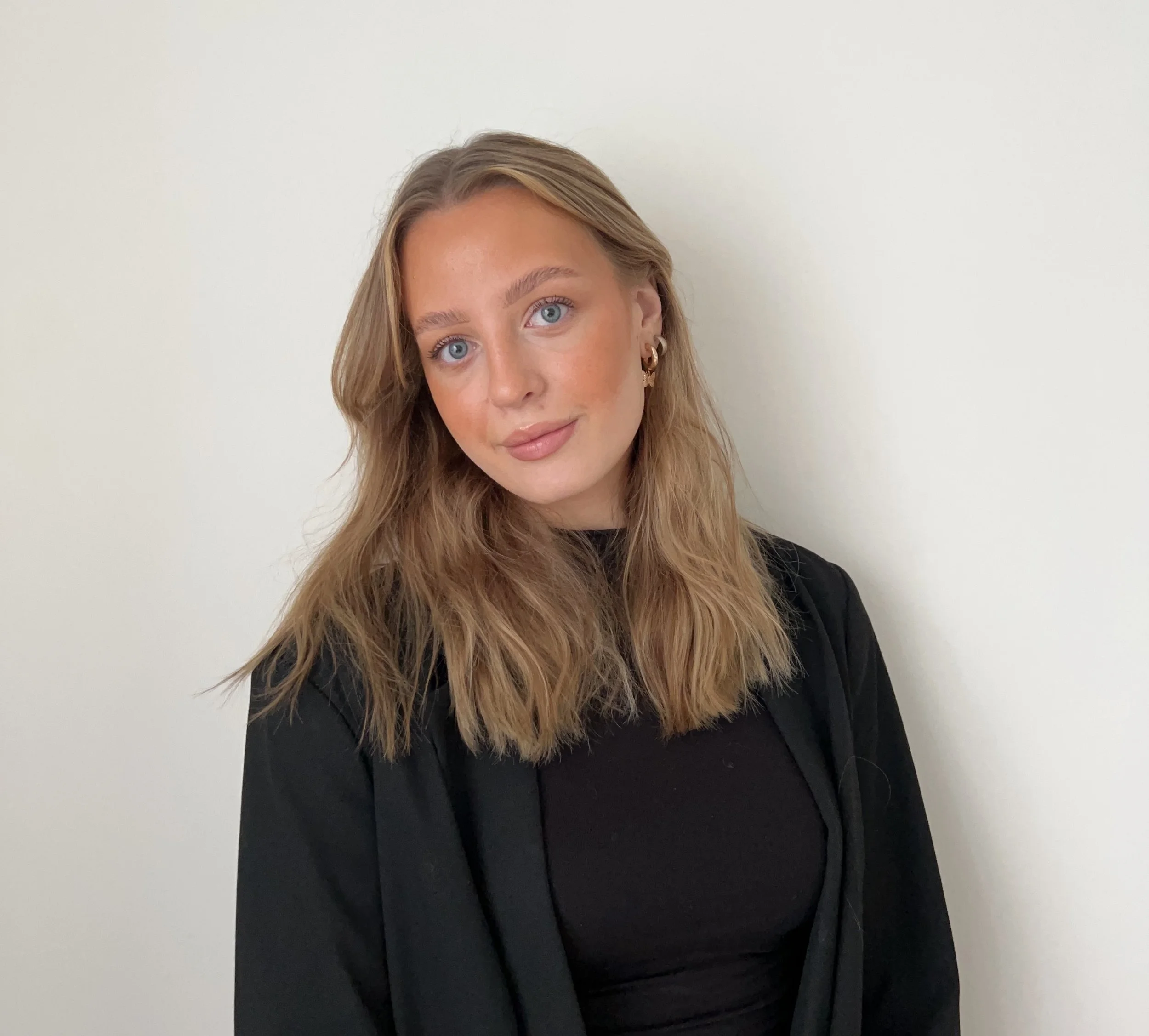 A woman with wavy, shoulder-length blonde hair and blue eyes posing against a plain white wall, wearing a black top and earrings.