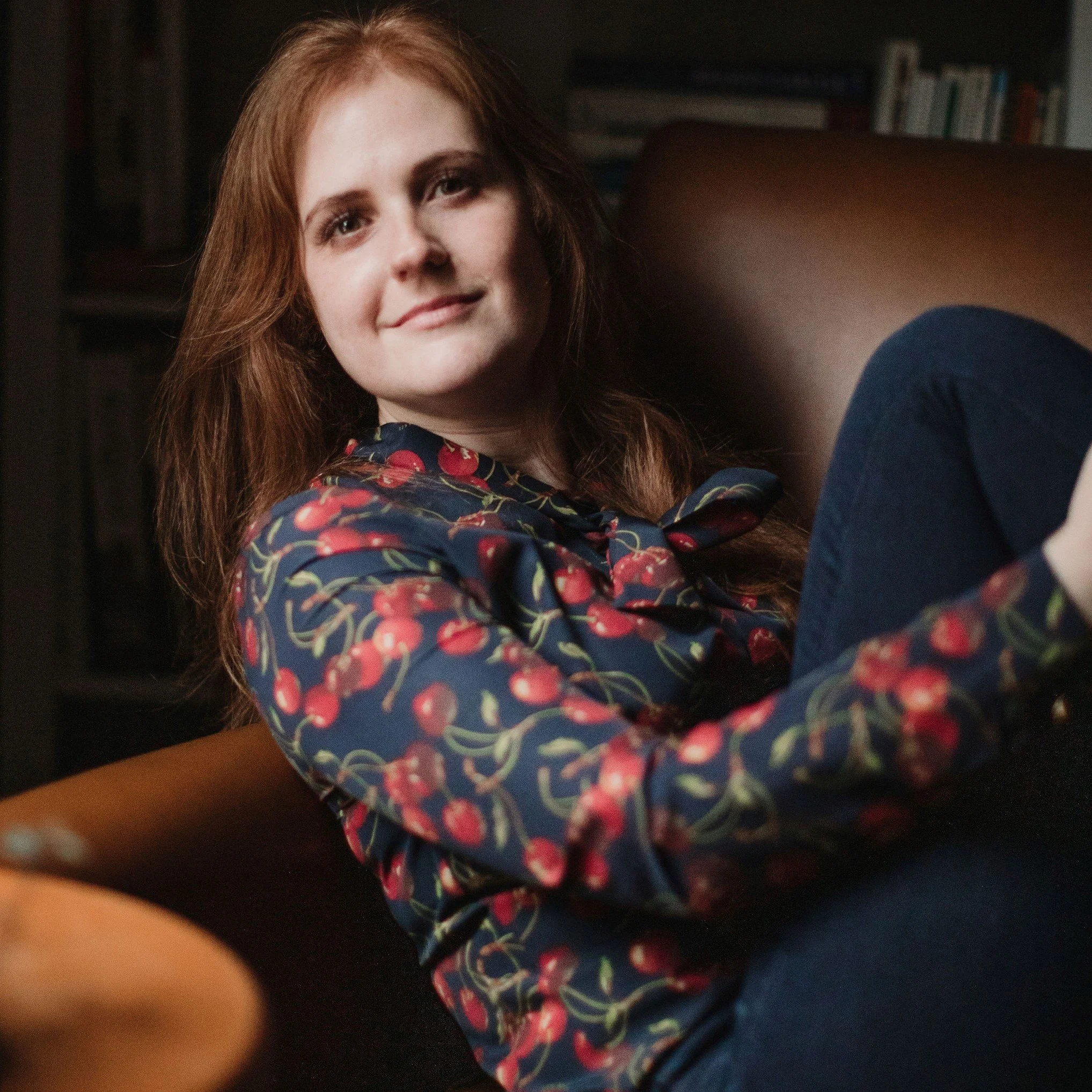Close-up of a woman with red hair smiling, wearing a dark top with a cherry pattern, in a dimly lit room with shelves in the background.