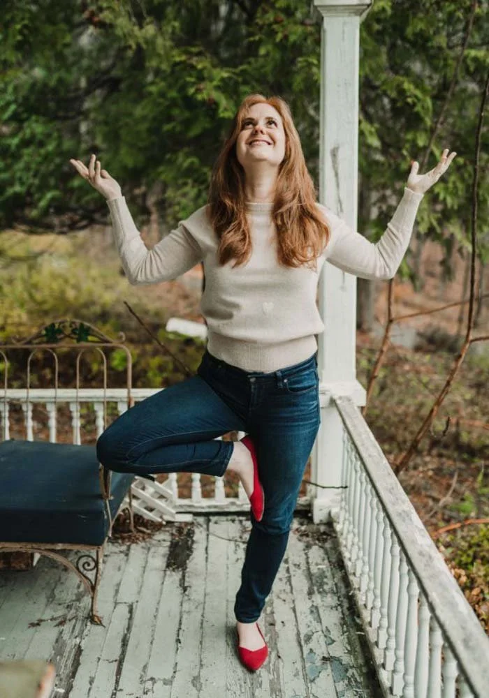 A woman with long red hair doing a yoga pose on her balcony, balancing on one leg with arms raised, wearing a beige sweater, dark jeans, and red heels, with trees and foliage in the background.