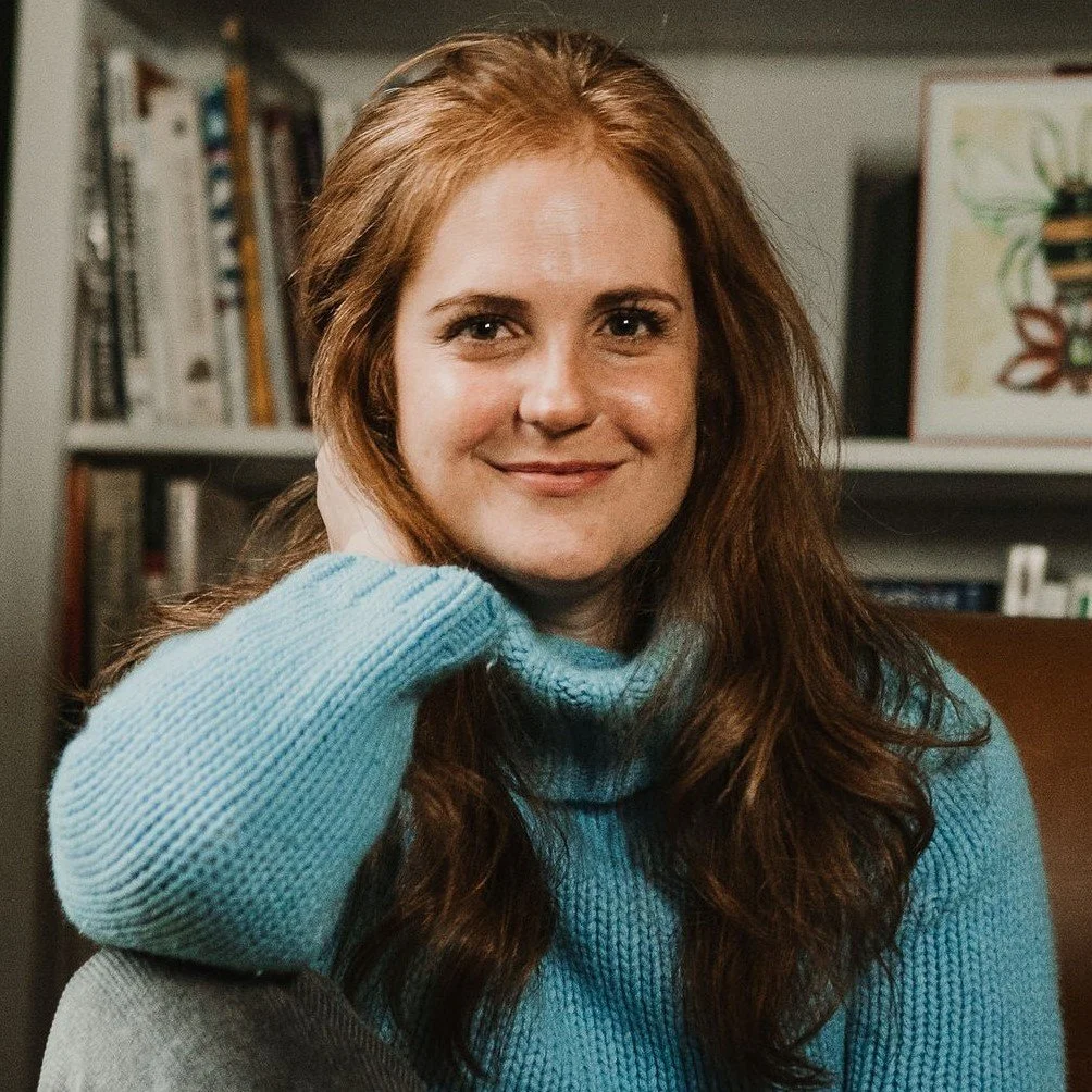 A woman with long, wavy, reddish-brown hair smiling at the camera, wearing a light blue turtleneck sweater, in a room with books and artwork in the background.