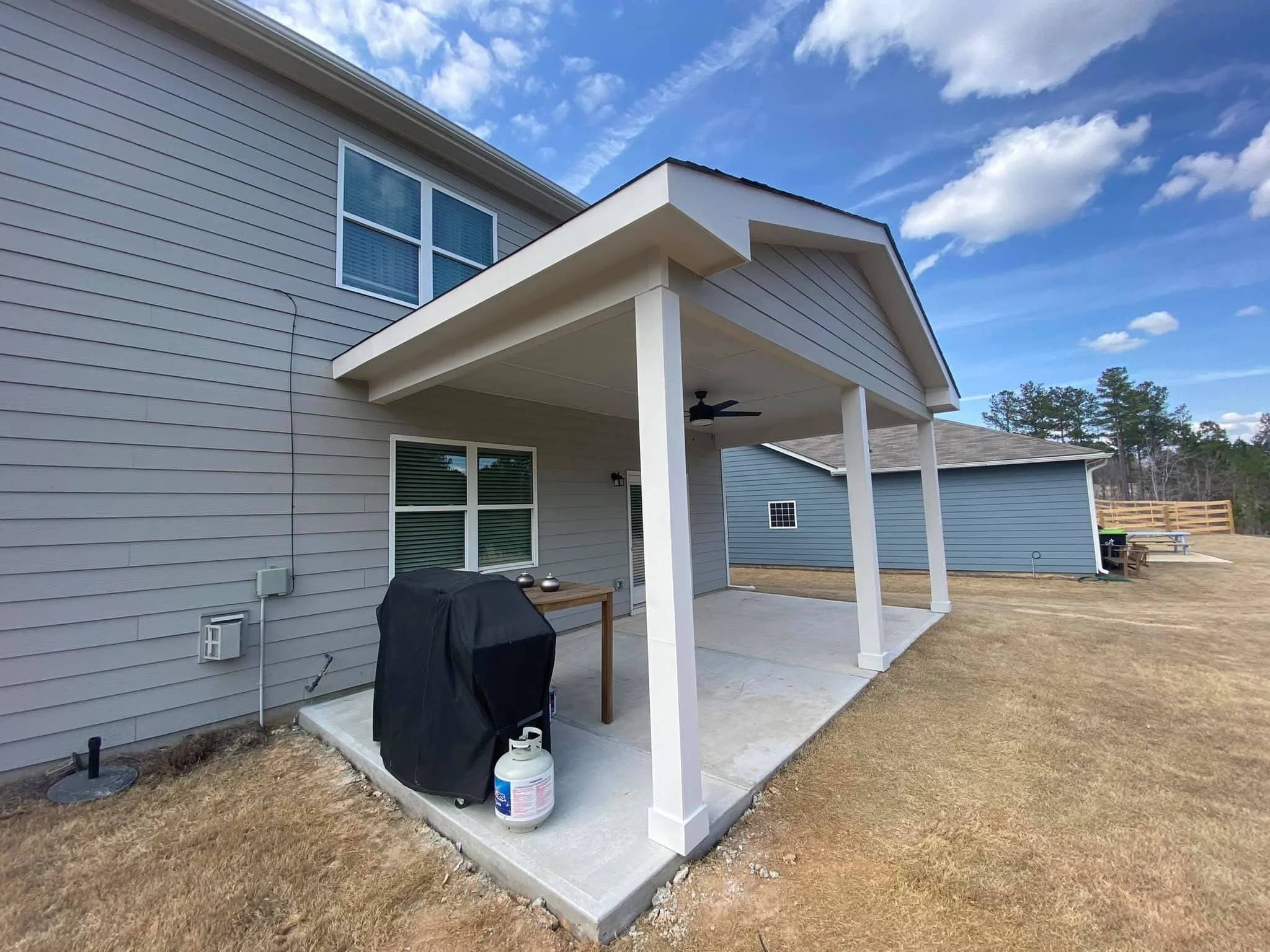 Backyard patio with concrete floor, contained by siding house, with a barbecue grill covered by a black cover, propane tank in front, and a small wooden table with decorative balls on top. To the right is a small grassy area with a picnic table, in a rural area with trees in the background and a partly cloudy sky.