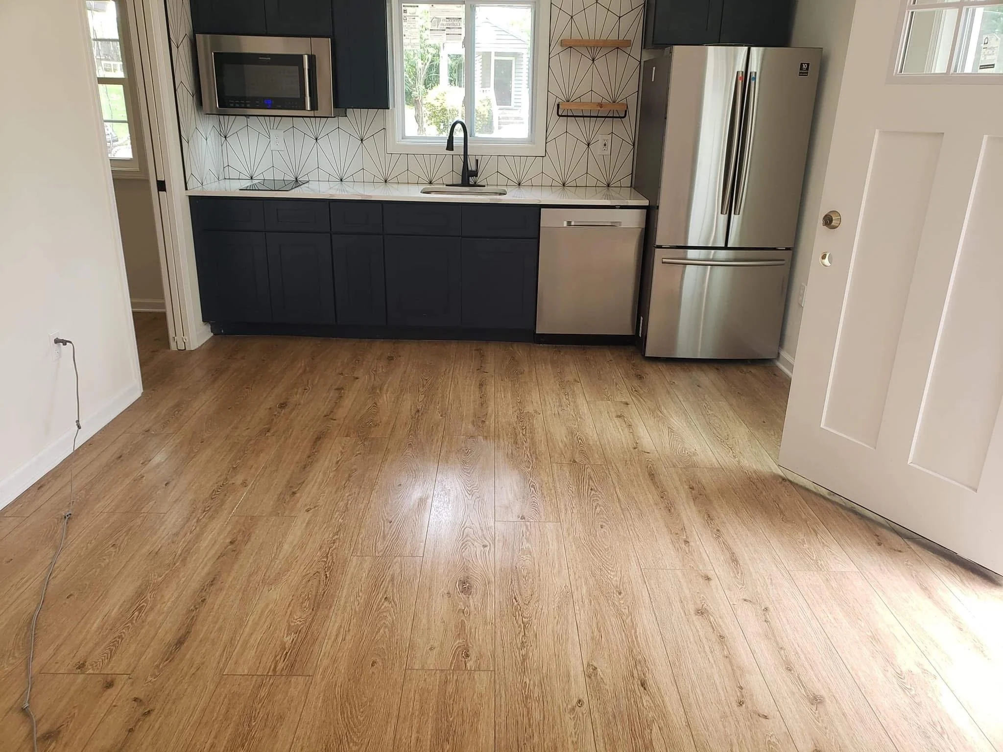 Modern kitchen with wood flooring, dark cabinets, stainless steel refrigerator and dishwasher, microwave oven, white countertop, geometric patterned backsplash, window above sink, and shelves on wall.