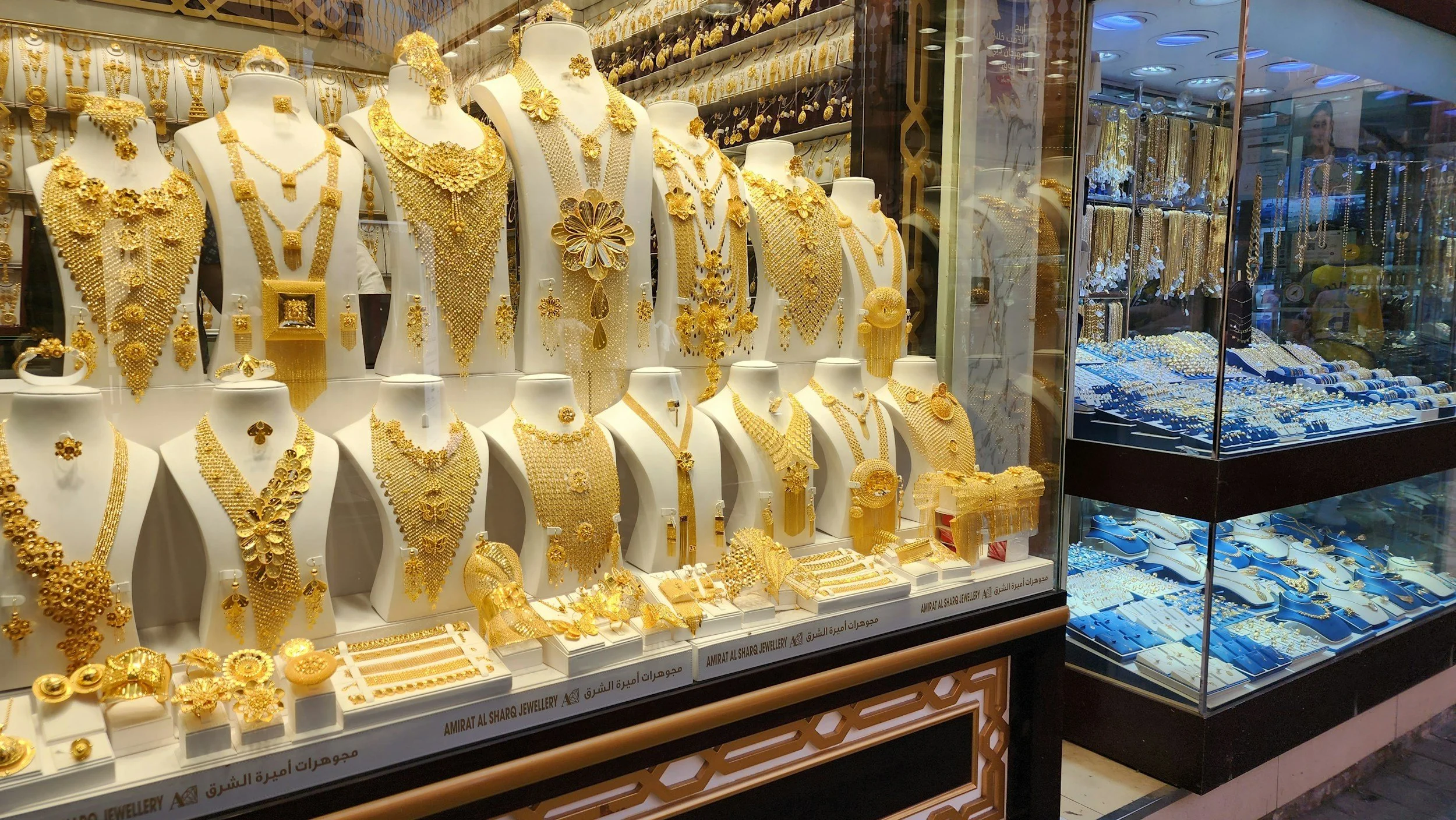 Display window of a jewelry store showcasing various gold jewelry including necklaces, rings, bracelets, and earrings, arranged on mannequin busts and trays, with additional jewelry visible in glass cases.