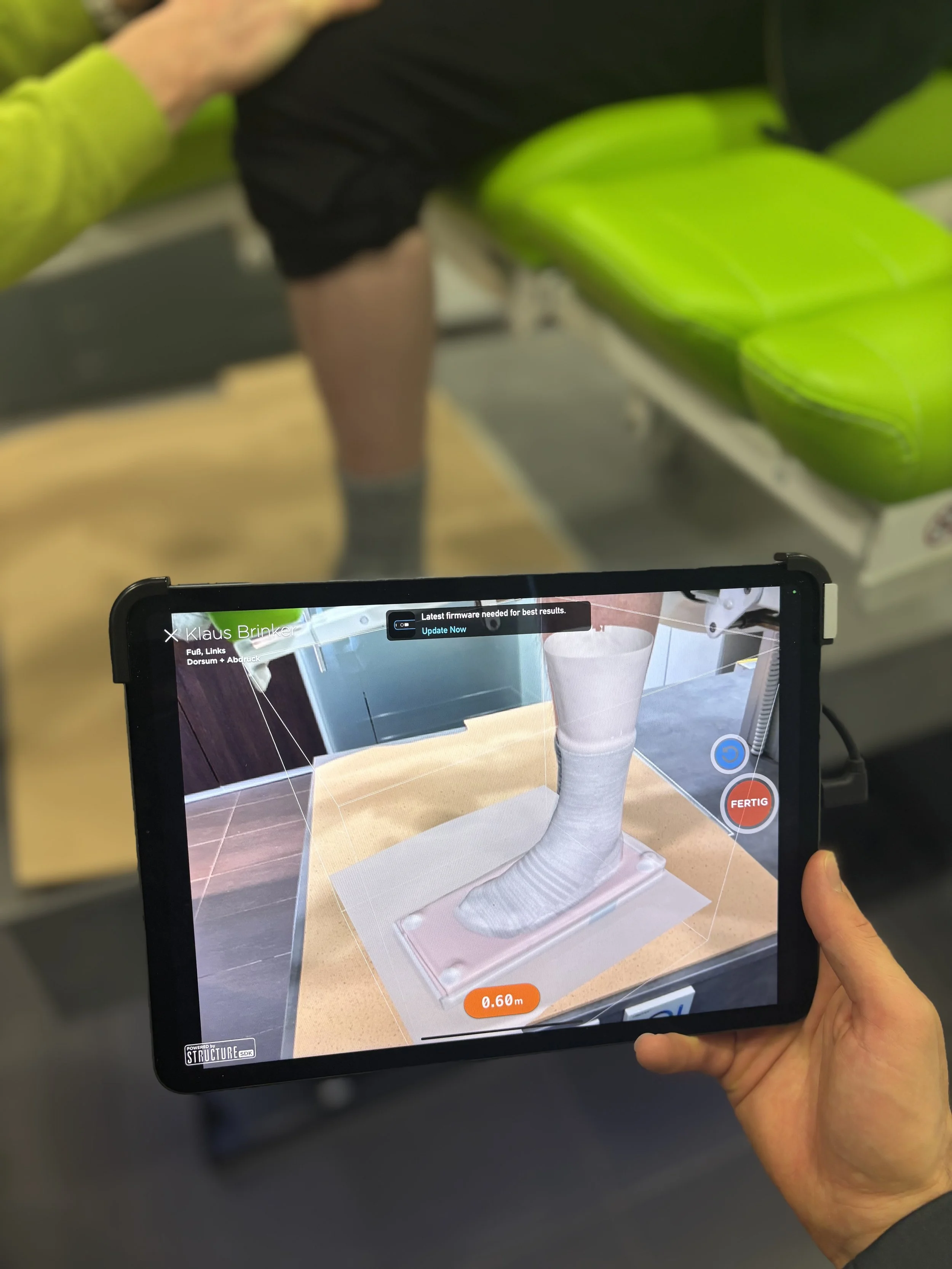 Ein Tablet zeigt eine 3D-Visualisierung eines Fußes, während im Hintergrund die Person sitzt desen Füße gerade gescannt worden sind