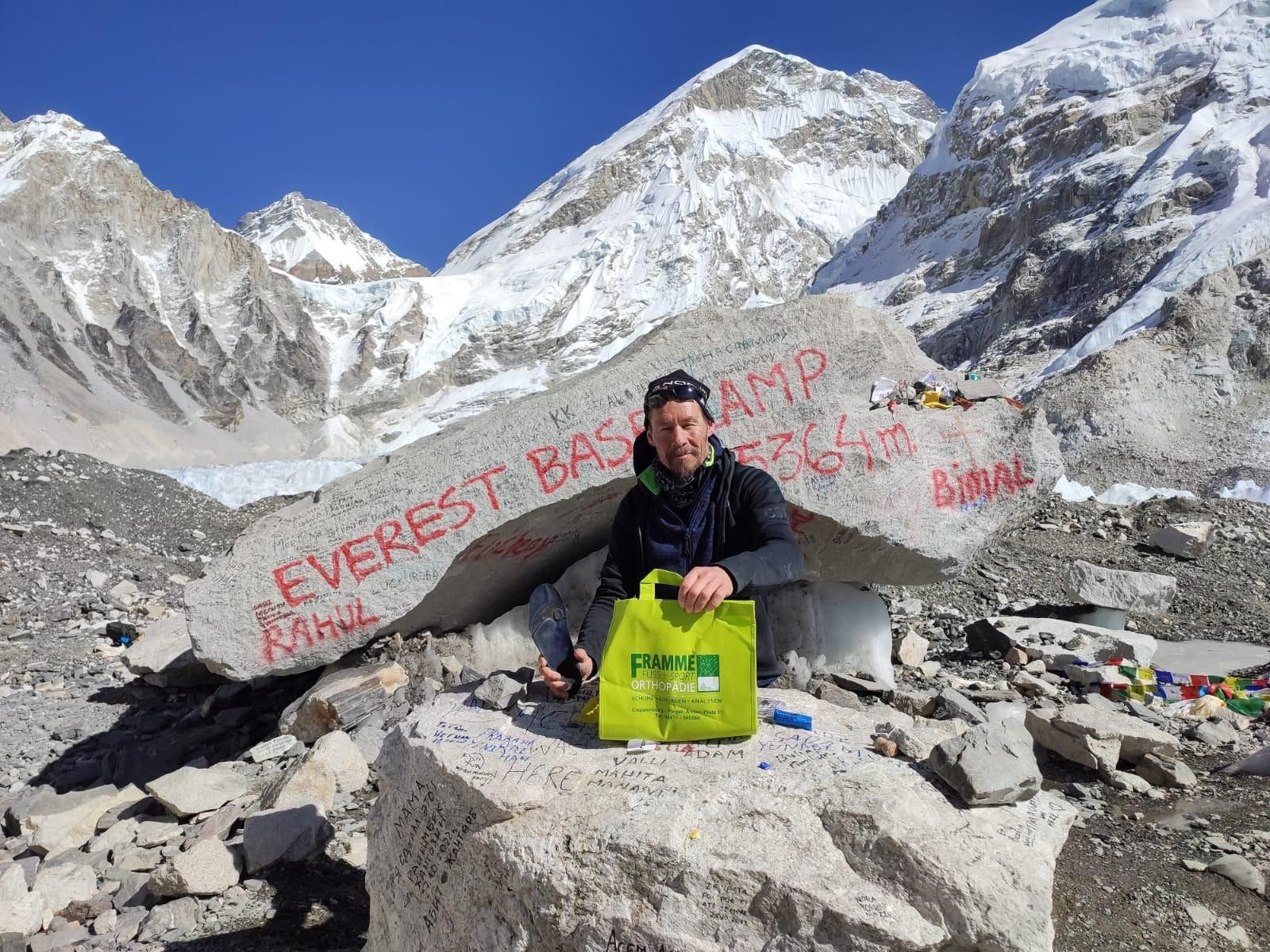 Ein Mann sitzt vor dem Check Point des Mount Everst Base Camps auf über 5000m Höhe und hält eine Framme Fuss+Sport Tasche und eine sensomotorische Einlage hoch