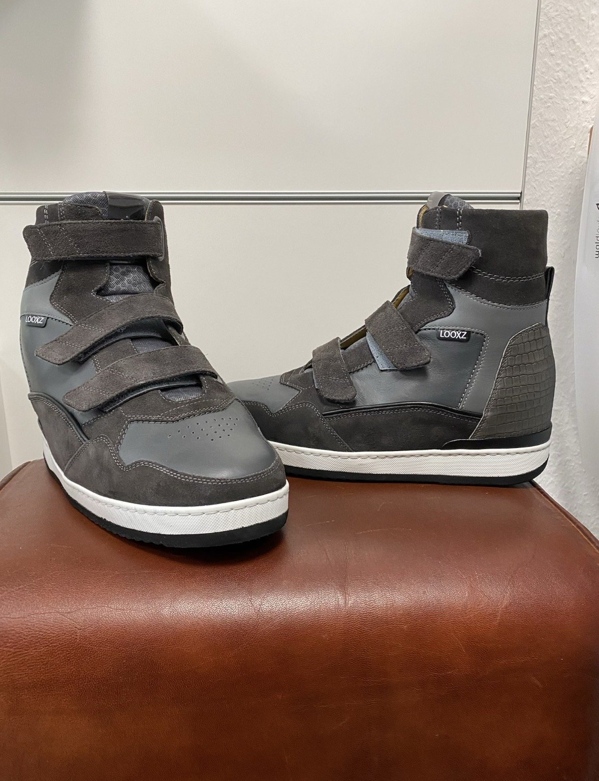 Ein Paar dunkle, hochtop Sneaker mit Velours- und Lederelementen, Klettverschlüssen und der Marke Looxz, auf braunem Lederhocker vor einer weißen Wand.