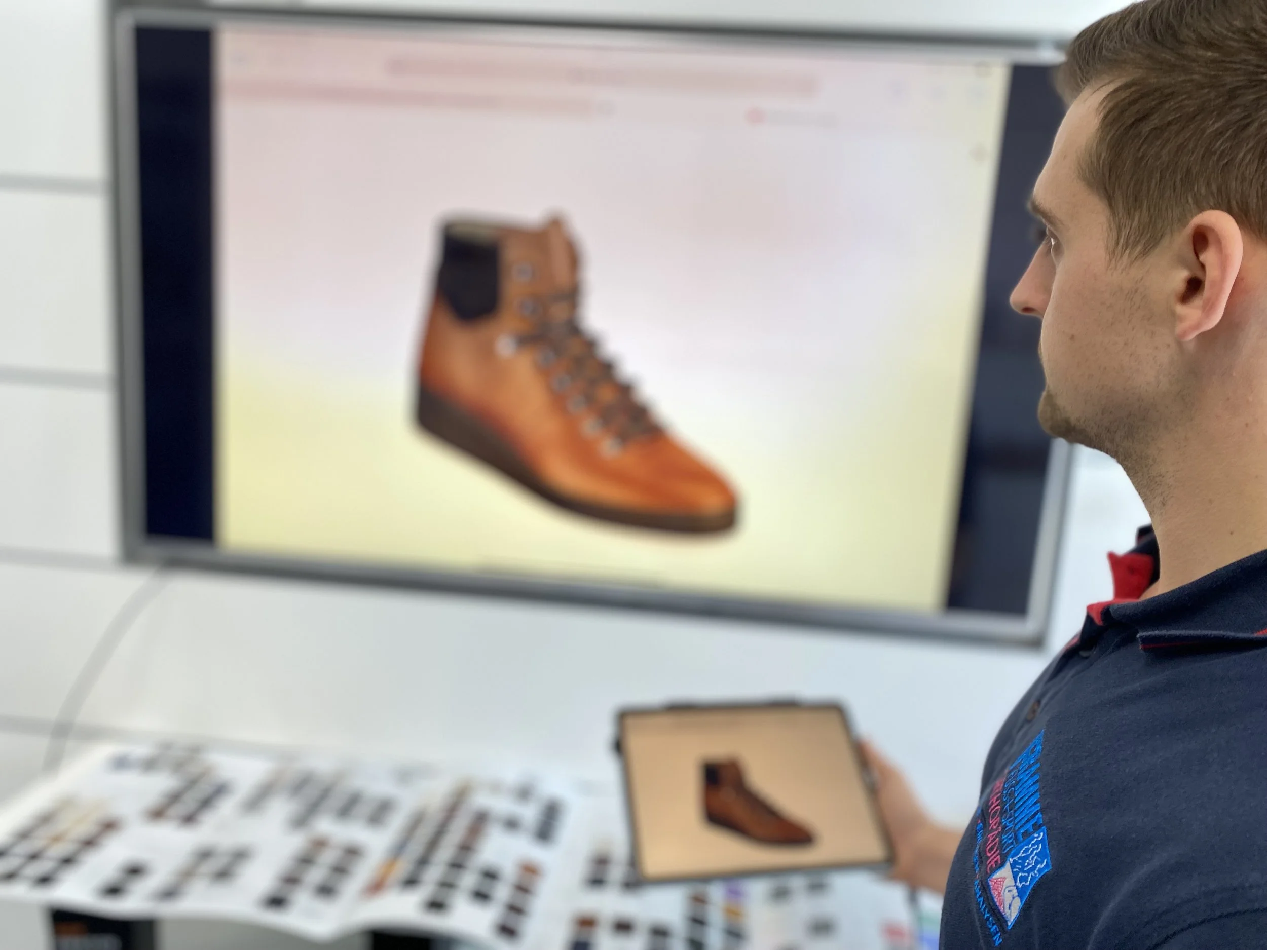 Ein Mann betrachtet ein digitales Abbild eines braunen Bergstiefels auf einem Bildschirm und hält ein Tablet mit einem ähnlichen Bild in der Hand. Hier wird das genaue Schuhmodell für den Maßschuh auf Kundenwunsch besprochen.