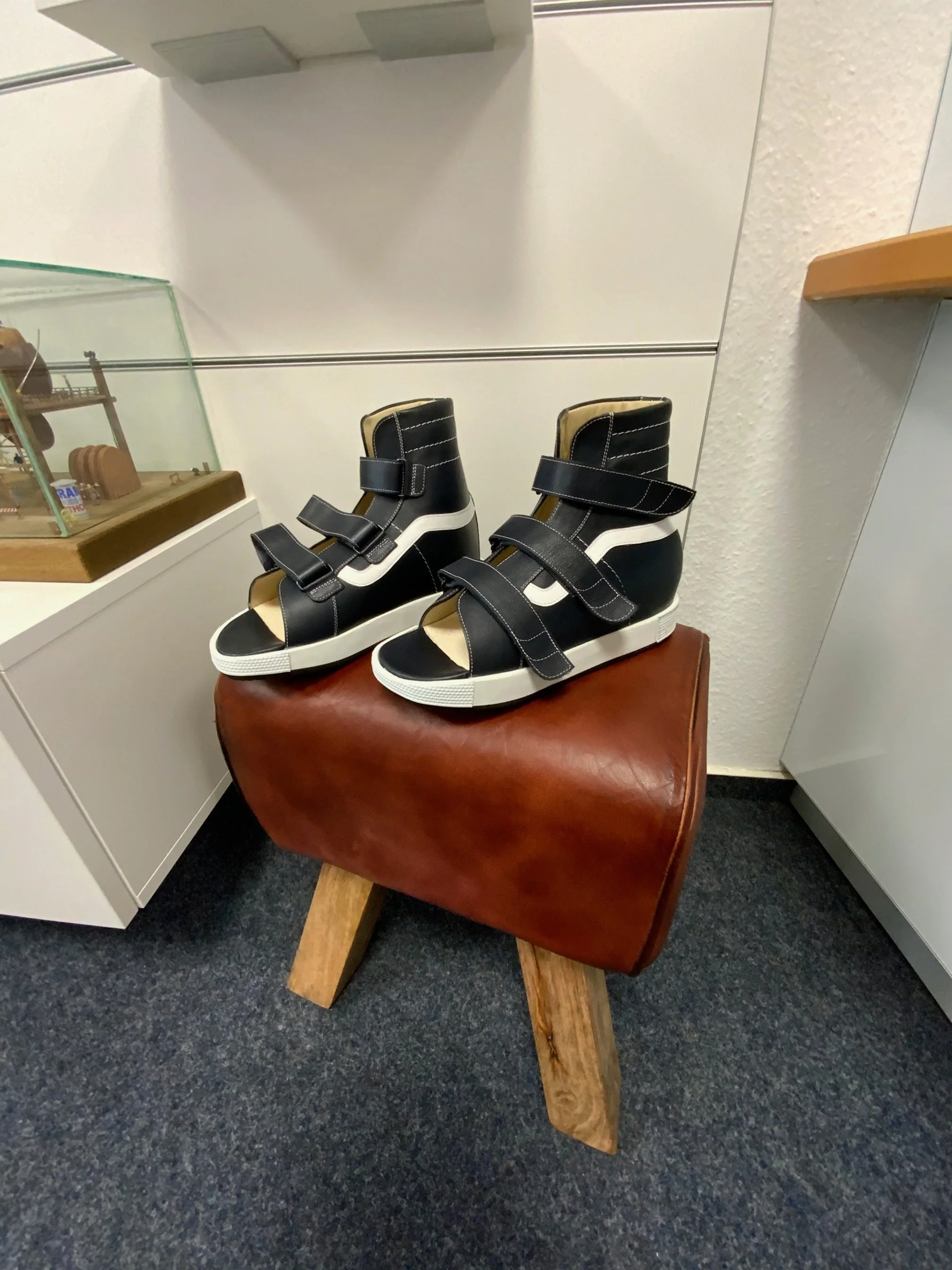 Schwarze und weiße hochgeschnittene Sandalen mit Klettverschluss stehen auf einem braunen Lederhocker.