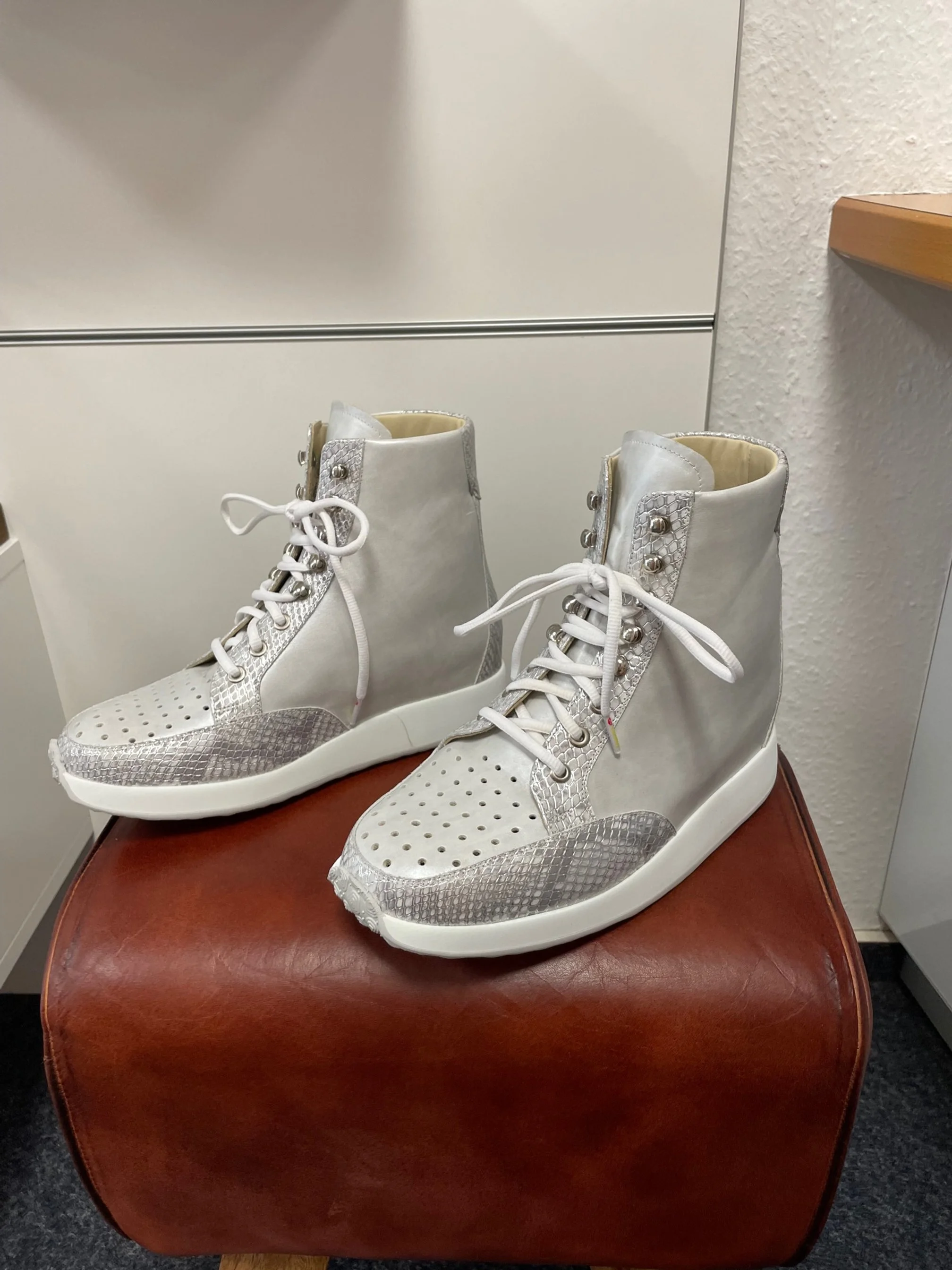 Weiße High-Top-Sneaker mit silbernen Details, Leder- und Snake-Print-Design, auf einem braunen Hocker in einem Raum.