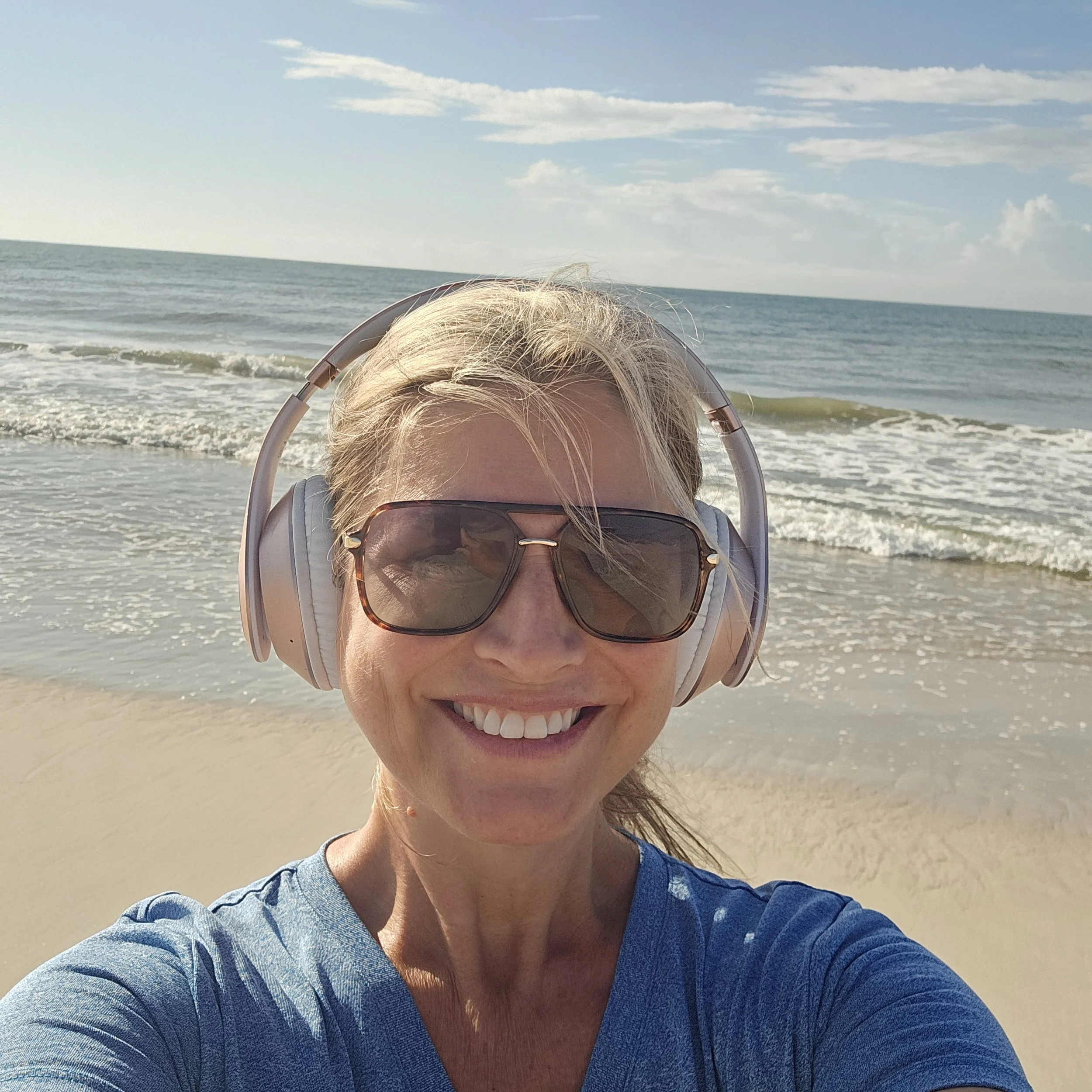 Woman smiling on the beach with sunglasses and headphones, ocean waves in the background.