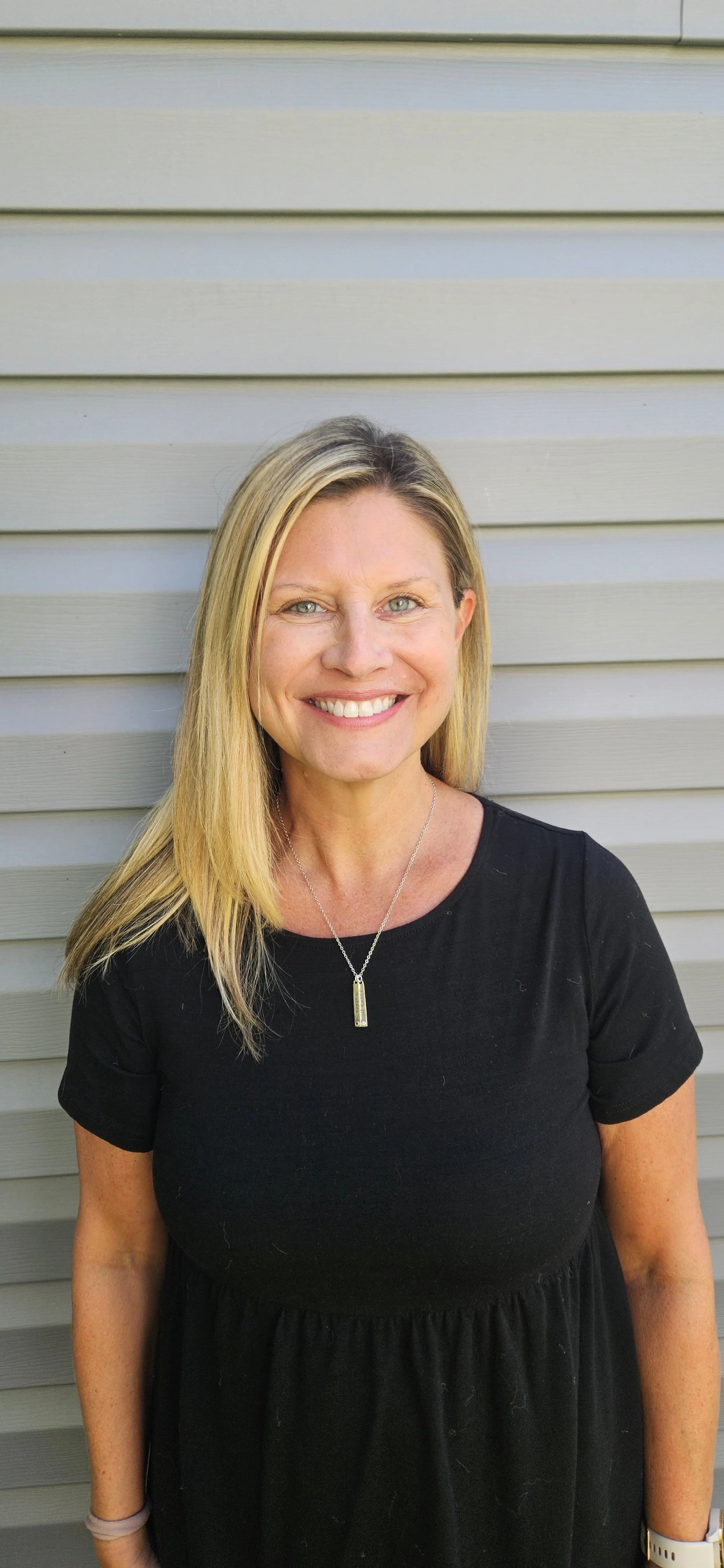 A smiling woman with blonde hair, wearing a black dress, necklace, and watch, standing in front of a light gray, horizontally-striped siding wall.