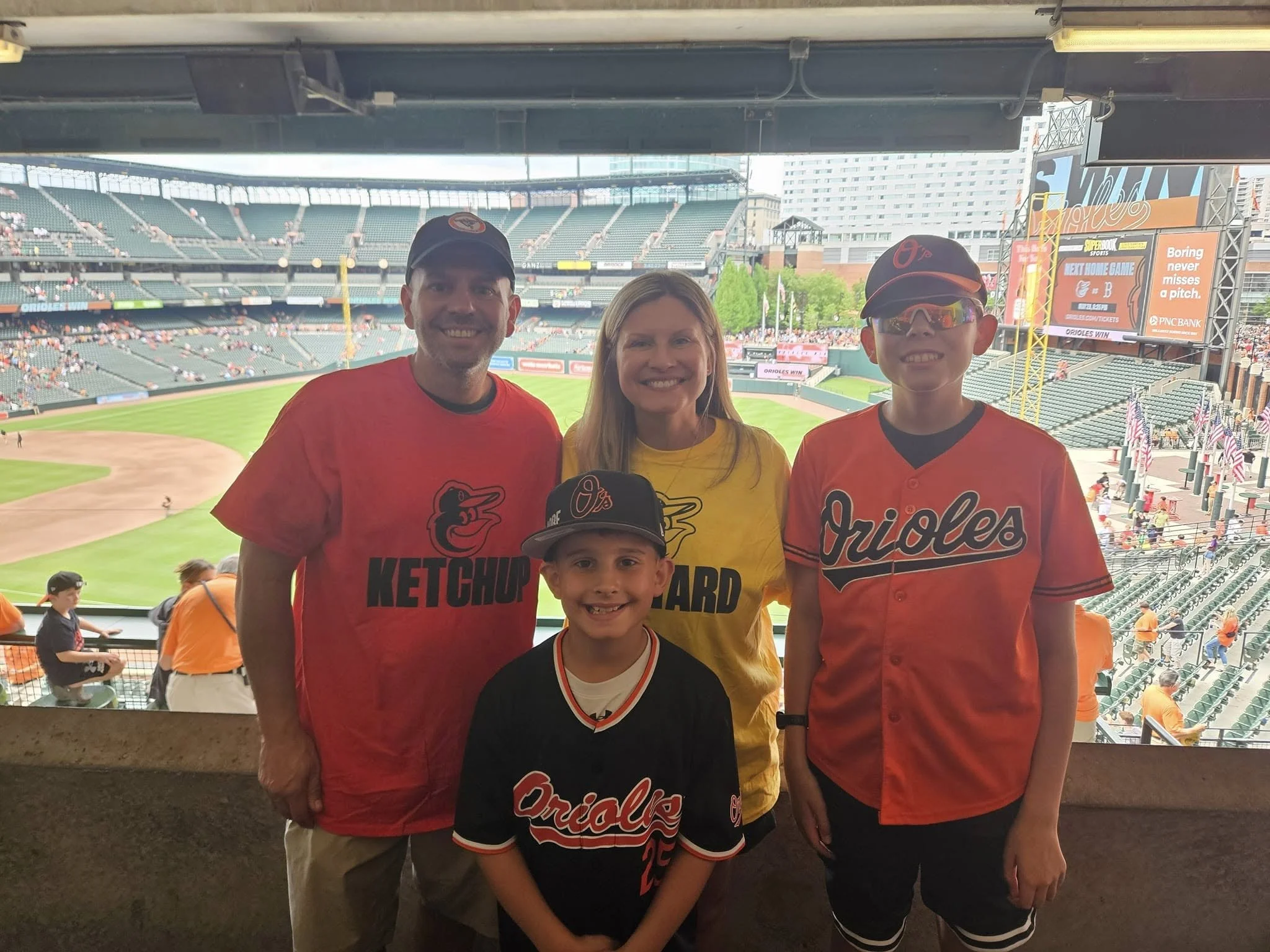Family of four at a baseball stadium, all wearing Baltimore Orioles team apparel, with the field in the background.