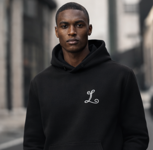 A young man wearing a black hoodie with a white embroidered letter 'L' on the left chest, standing outdoors in an urban setting with blurred buildings in the background.