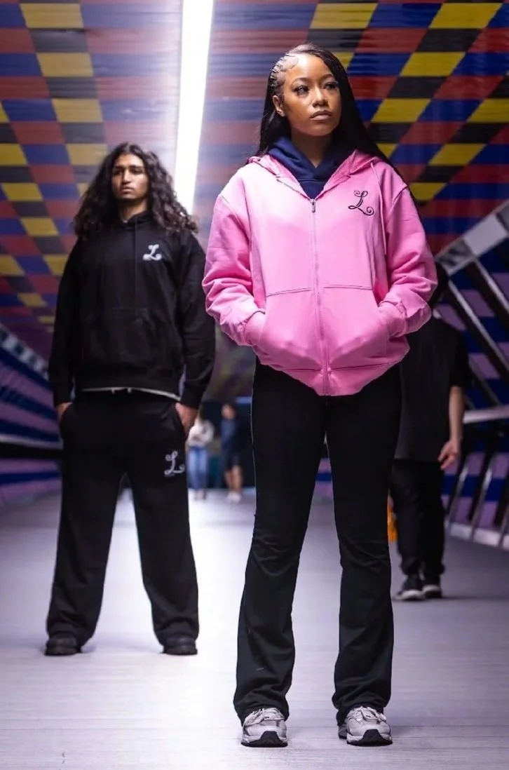 Two women standing in a colorful tunnel, wearing matching hoodies and sweatpants with a logo, with other people in the background.