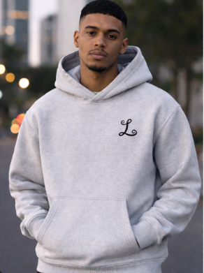 Young man in a gray hoodie with an embroidered letter 'L' standing outdoors at dusk.