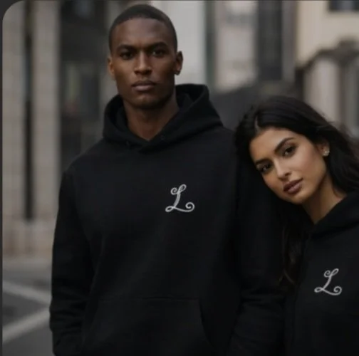 A young man and woman standing outdoors near buildings, both wearing black hoodies with a white 'L' logo.