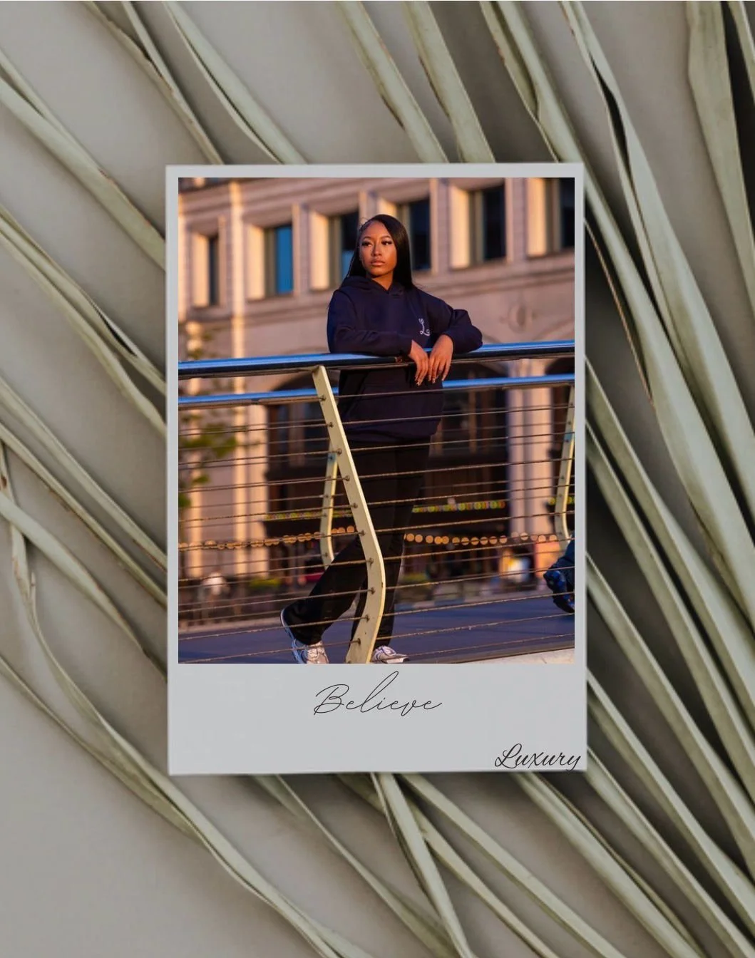 A young woman with long dark hair leaning on a city bridge railing at sunset, wearing a dark hoodie and black pants, with a building in the background. The photo has a caption that reads "Believe" and "Luxury."