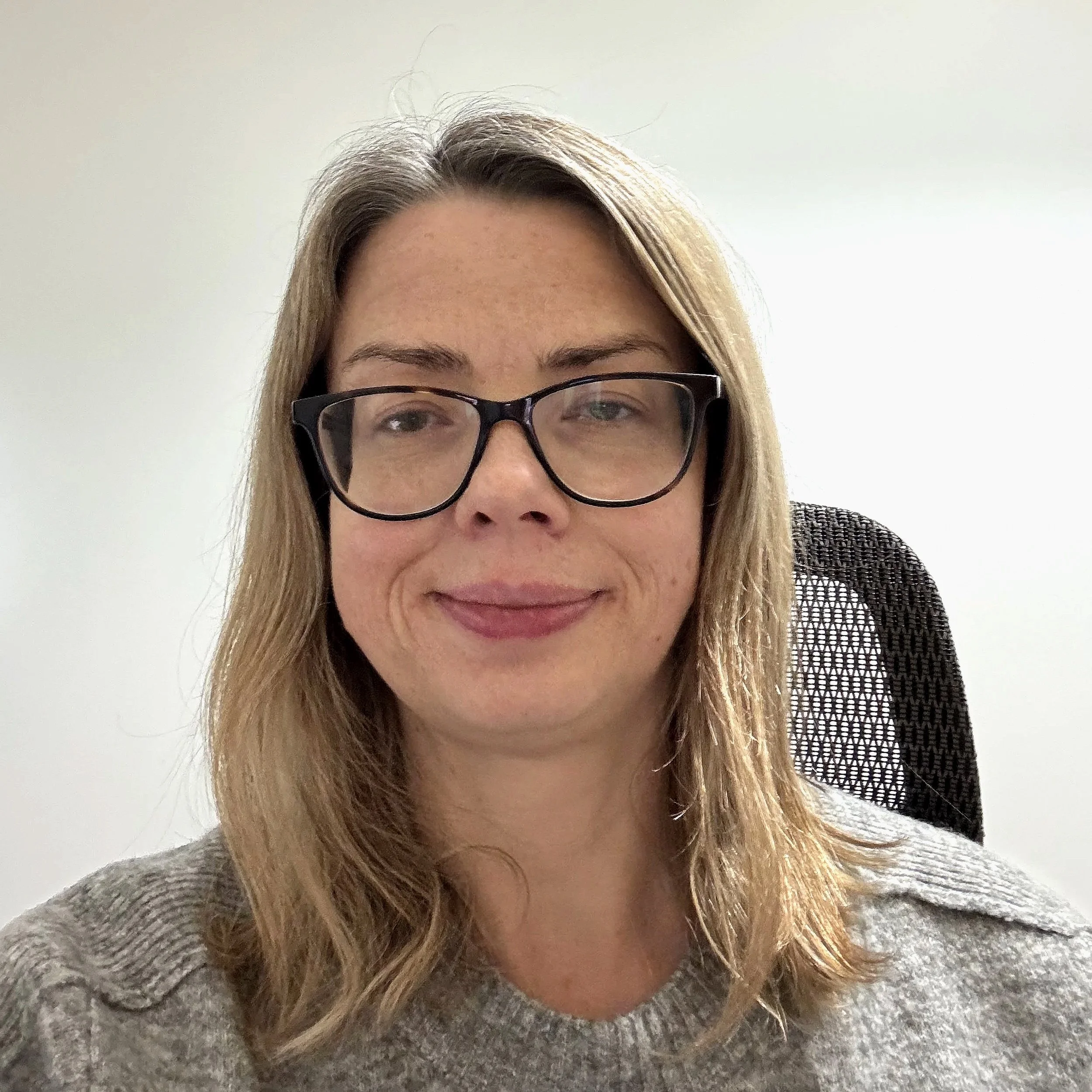 A woman with long blonde hair, wearing glasses, sitting in a gray sweater, smiling at the camera, with a black office chair and plain white wall in the background.