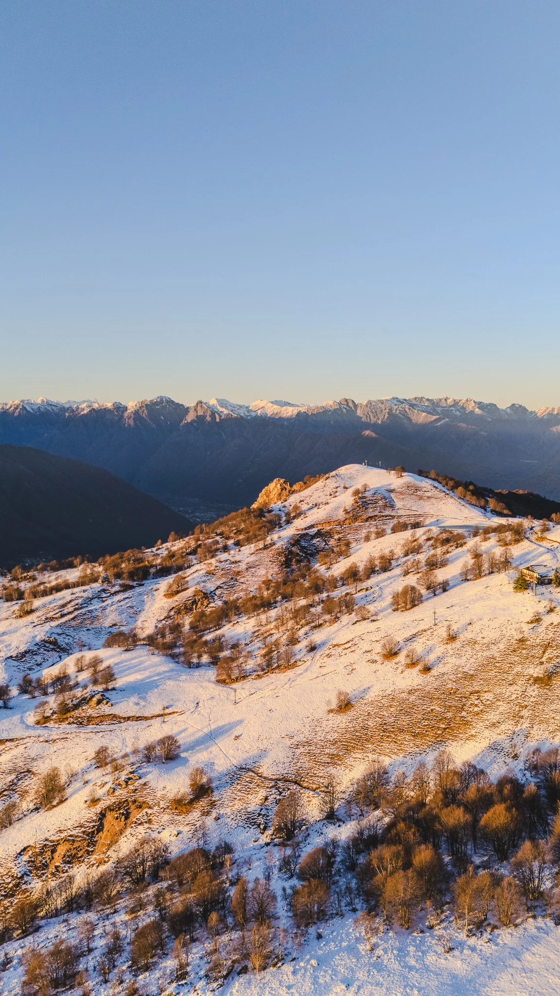 Paesaggio di montagna innevata con colline e alberi, con una catena montuosa sullo sfondo durante il tramonto.