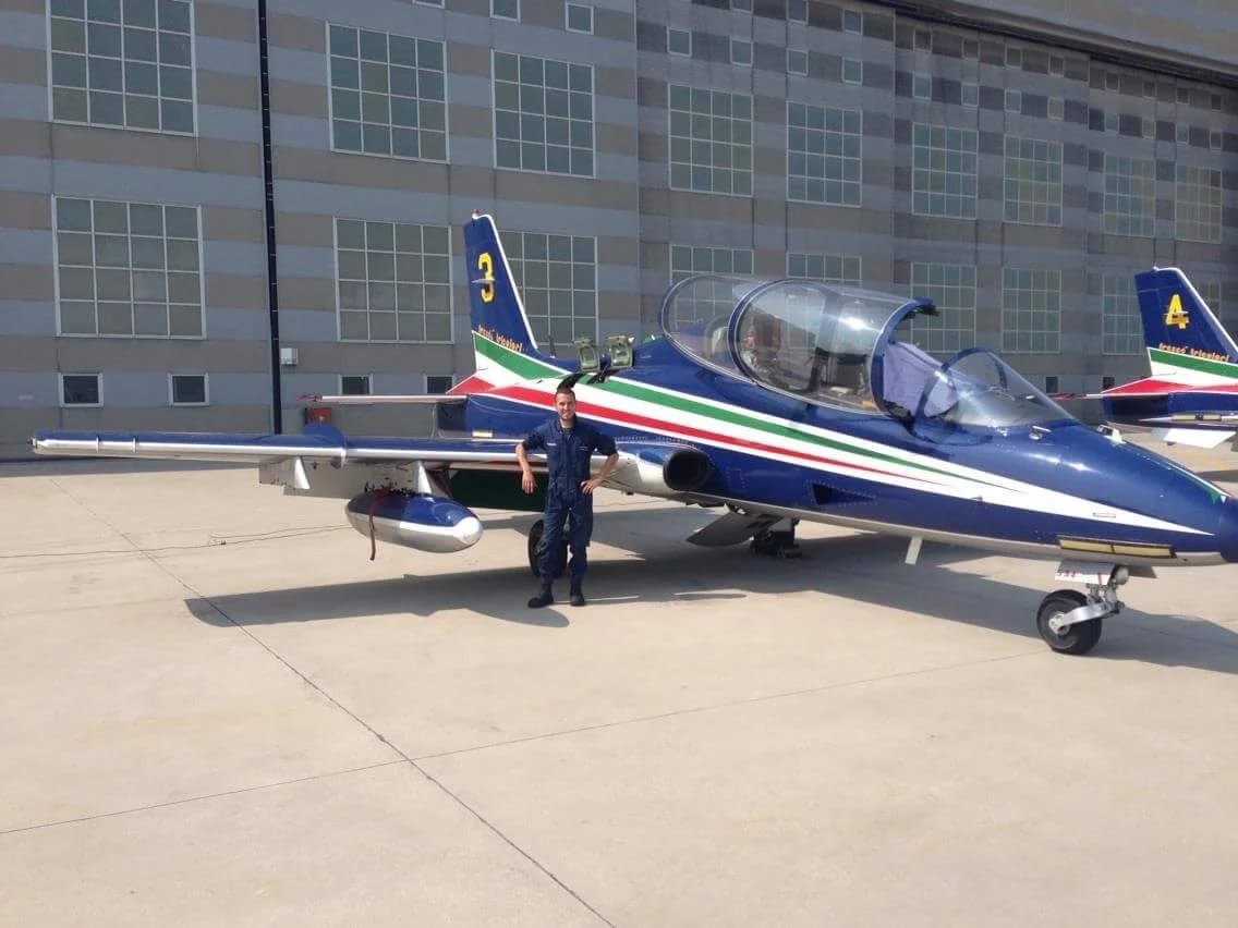 Un uomo in uniforme si trova davanti a un aereo da combattimento colorato con il tricolore italiano, parcheggiato su un'area di cemento con un edificio sullo sfondo.